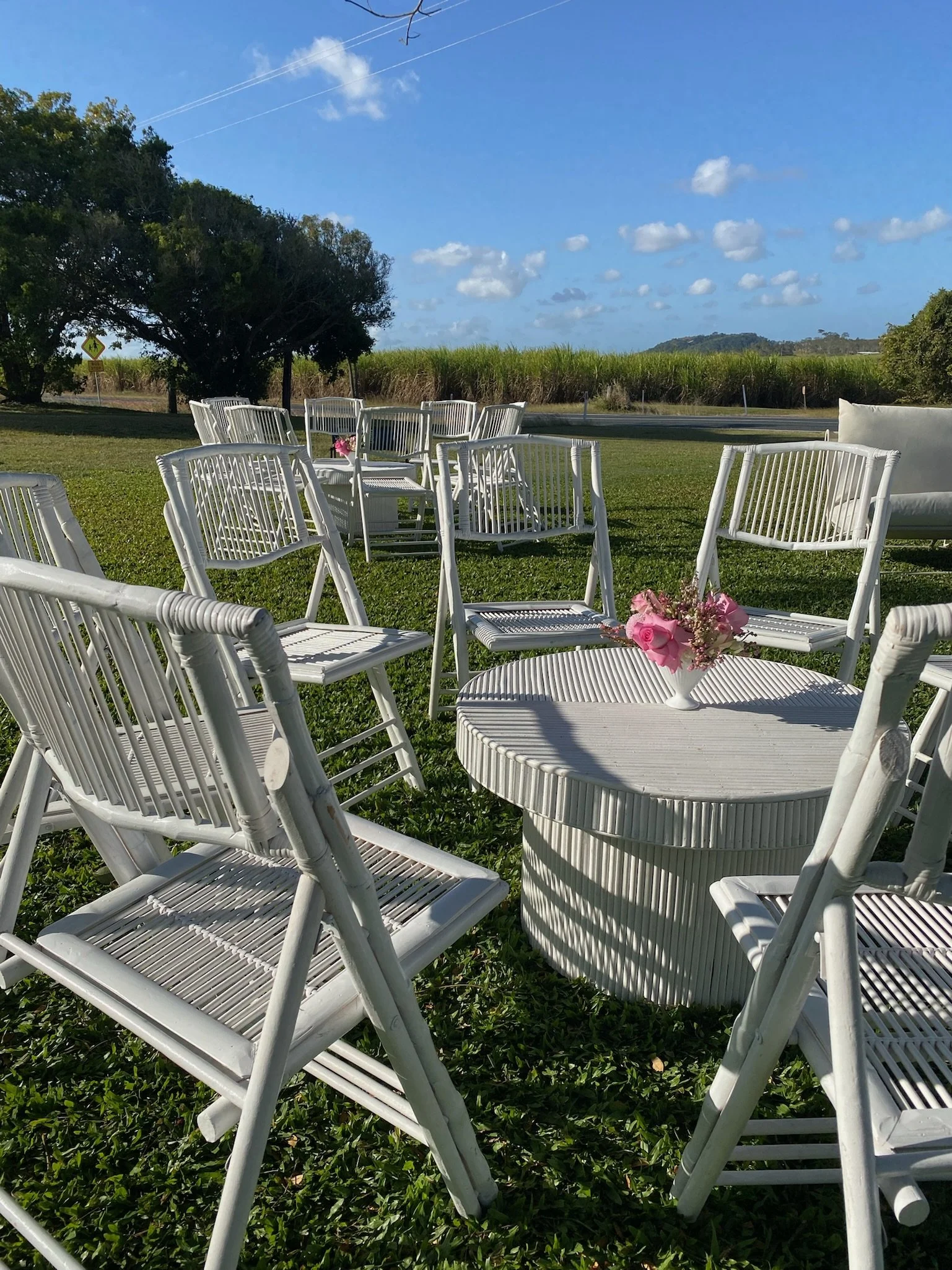 Outdoor wedding or event setup with white chairs arranged around a white, round table with pink flowers, on a grassy lawn with trees, a road, and a blue sky with clouds in the background.