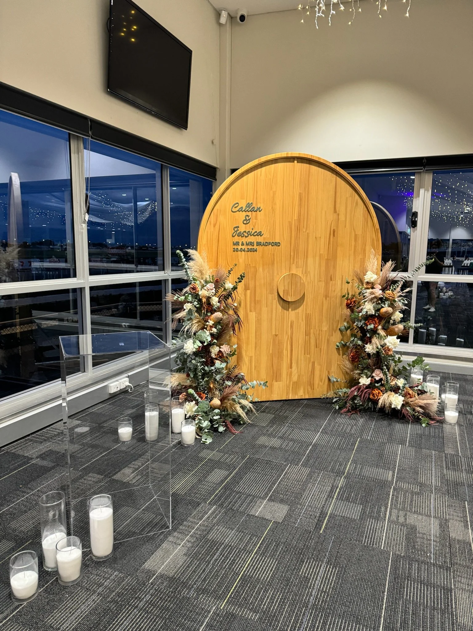 Wedding backdrop with wooden arch, floral arrangements, candles, and the names Callan and Jessica along with the date 20.04.2024, inside a venue with large windows, a TV on the wall, and string lights.