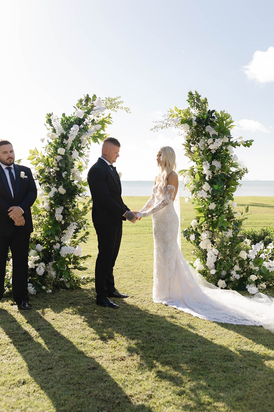 A wedding ceremony taking place outdoors on a grassy area near water. A bride and groom are holding hands and exchanging vows under a floral arch made of white flowers and green foliage. Two groomsmen in suits stand nearby, and the sky is bright with