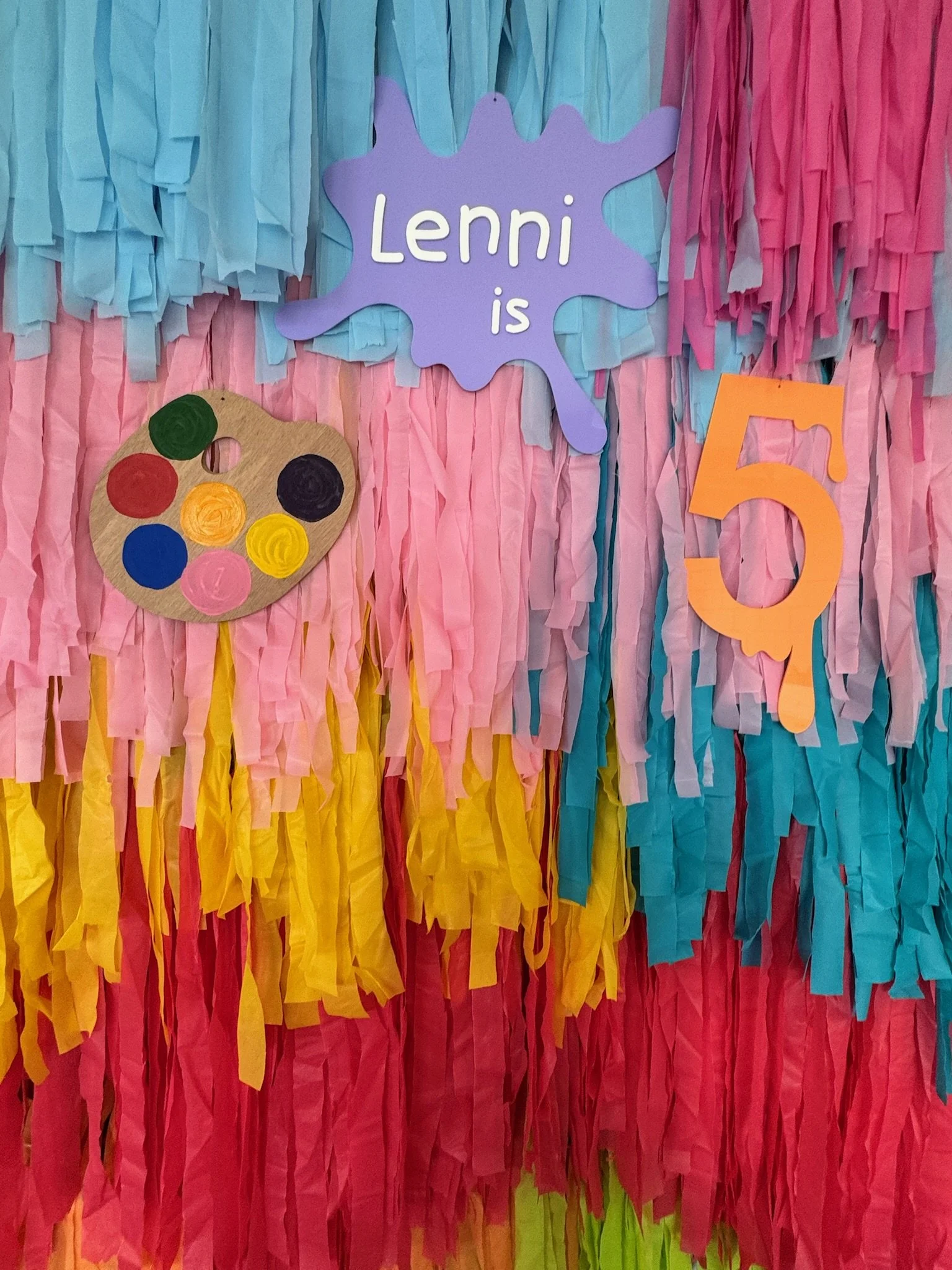 Colorful birthday decoration with tissue paper streamers in blue, pink, yellow, and red. Features a purple splash-shaped sign that says "Lenni is" and an orange cutout number 5. There is also a paper artist palette with colored paint dots.