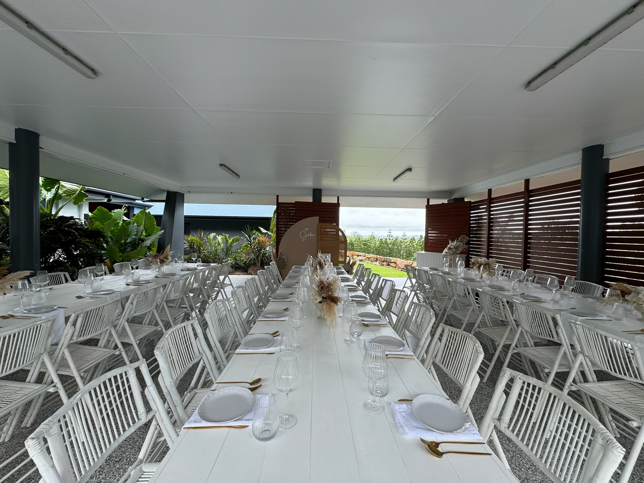 Long dining table set for a formal event with white chairs, plates, glasses, and gold utensils in an outdoor covered area with greenery and a view of the sky.