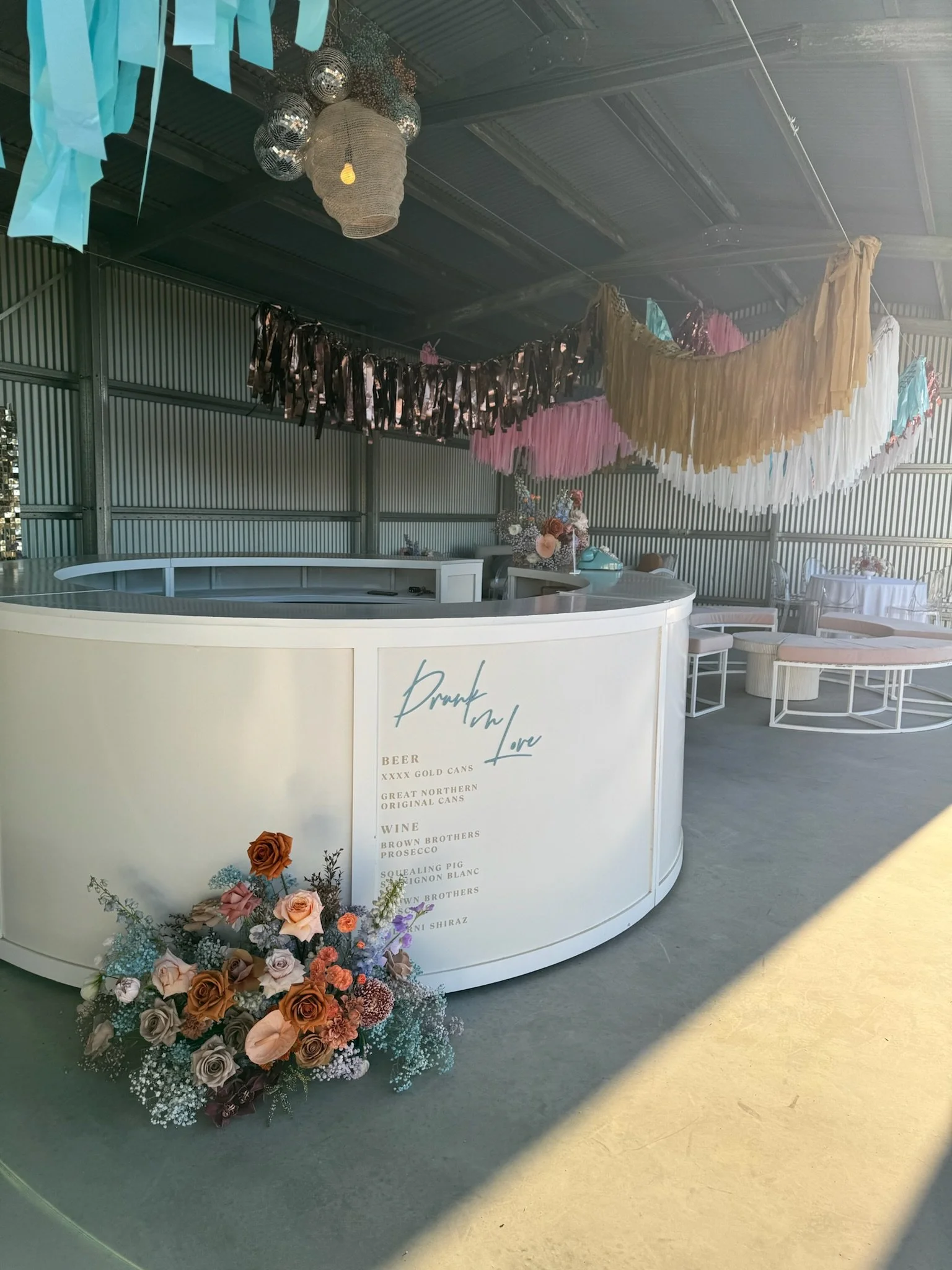 Decorated event space with pastel-colored tassel garlands hanging from the ceiling, a curved white bar with drink menu, floral arrangements with roses and baby's breath, and tables set up for guests.