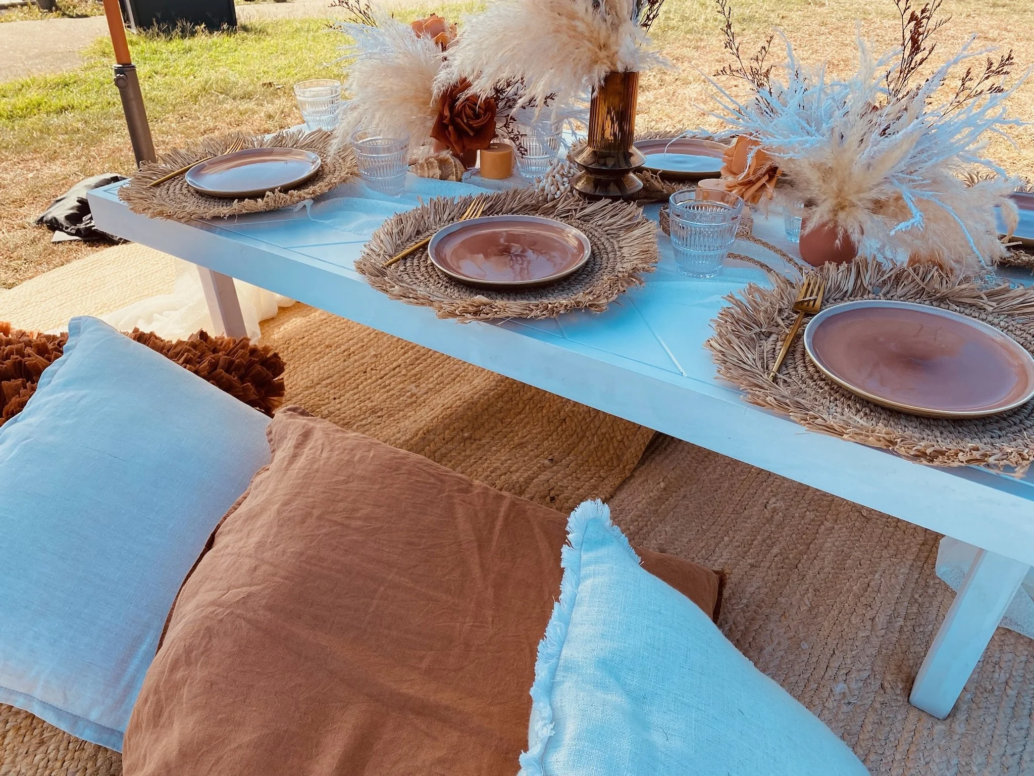 Table set for an outdoor meal with woven placemats, pink plates, clear glasses, and napkins with floral arrangements and decorative feathers.