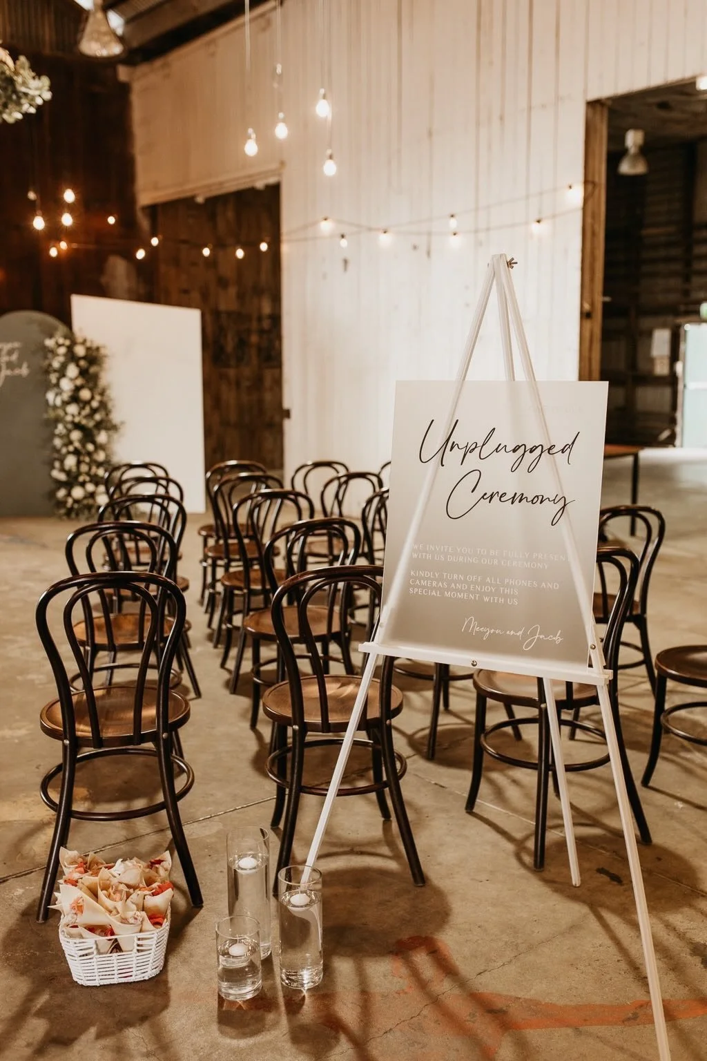 Indoor wedding ceremony setup with rows of chairs, floral arrangements, lit candles, and a sign that reads "Unplugged Ceremony" inviting guests to turn off phones and cameras.