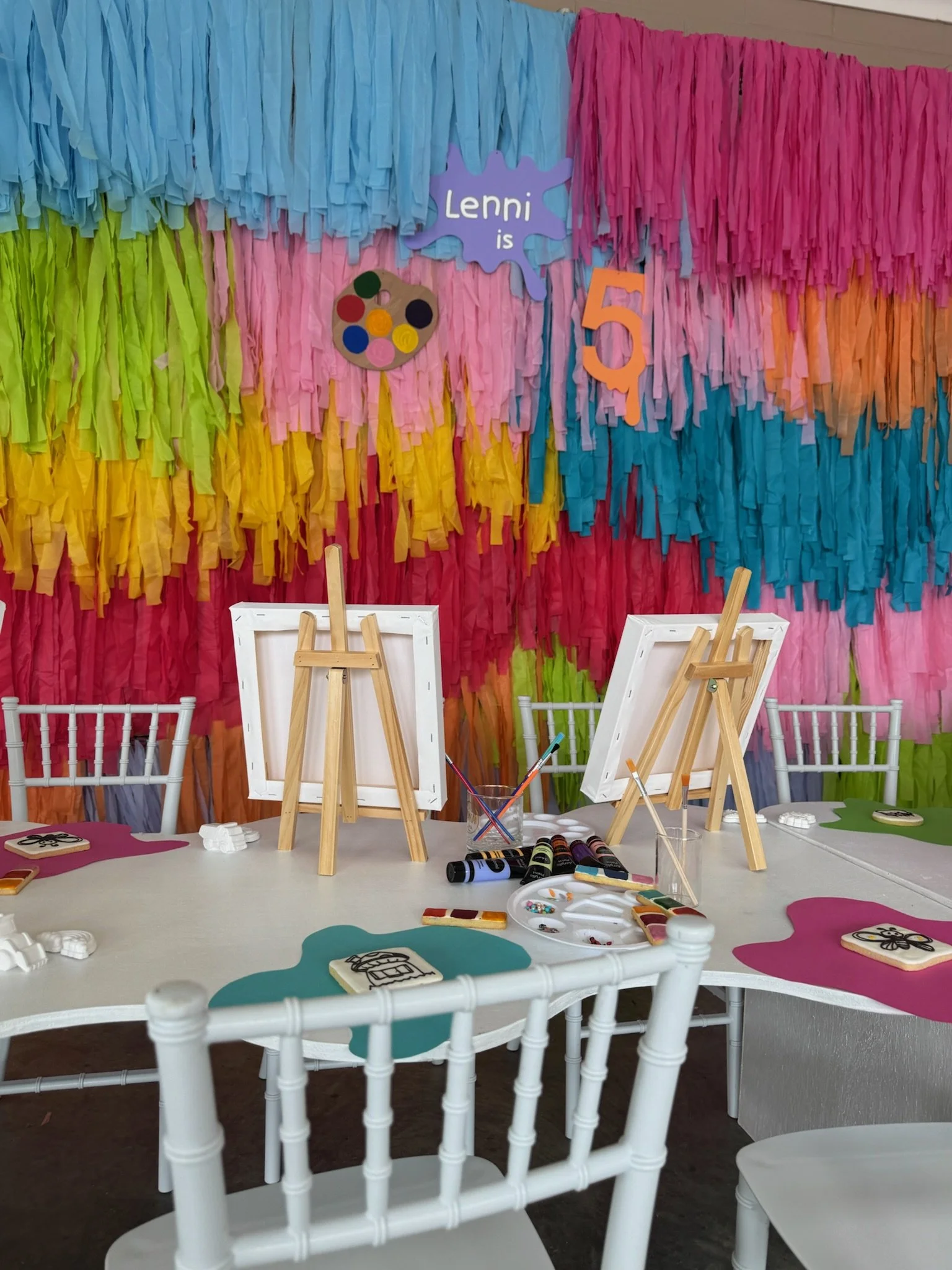 A colorful party backdrop with multicolored paper fringes, indicating a celebration for a child's fifth birthday, with a table set up for painting art activities.