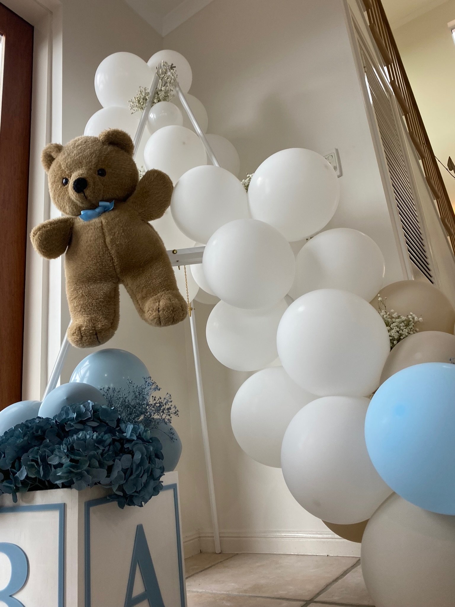 Decorative display with white and light blue balloons, blue flowers, a plush teddy bear with a blue bow tie, and a sign with partially visible letters.