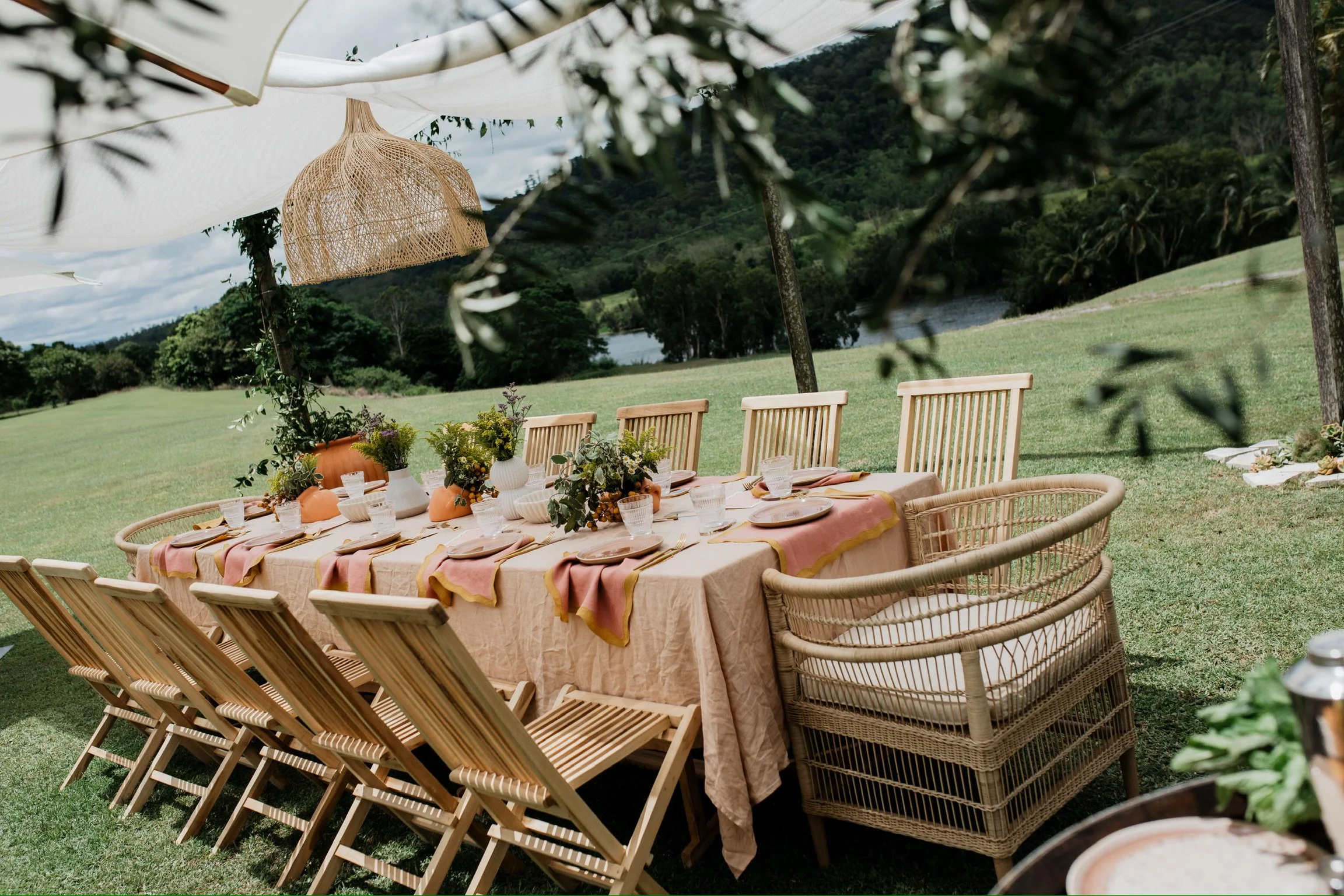 Outdoor dining table set up on a grassy field with a scenic landscape including trees, a lake, and hills in the background. The table has a beige tablecloth, pink and orange napkins, and is decorated with vases of flowers. Wicker chairs surround the 