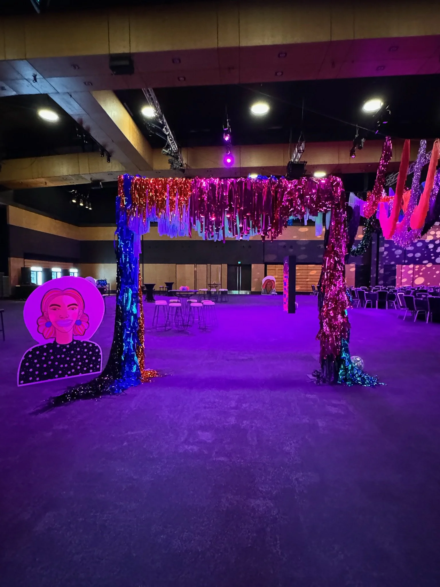 Pink and purple metallic hanging decorations cover an archway in a large, empty event space with stage and seating.