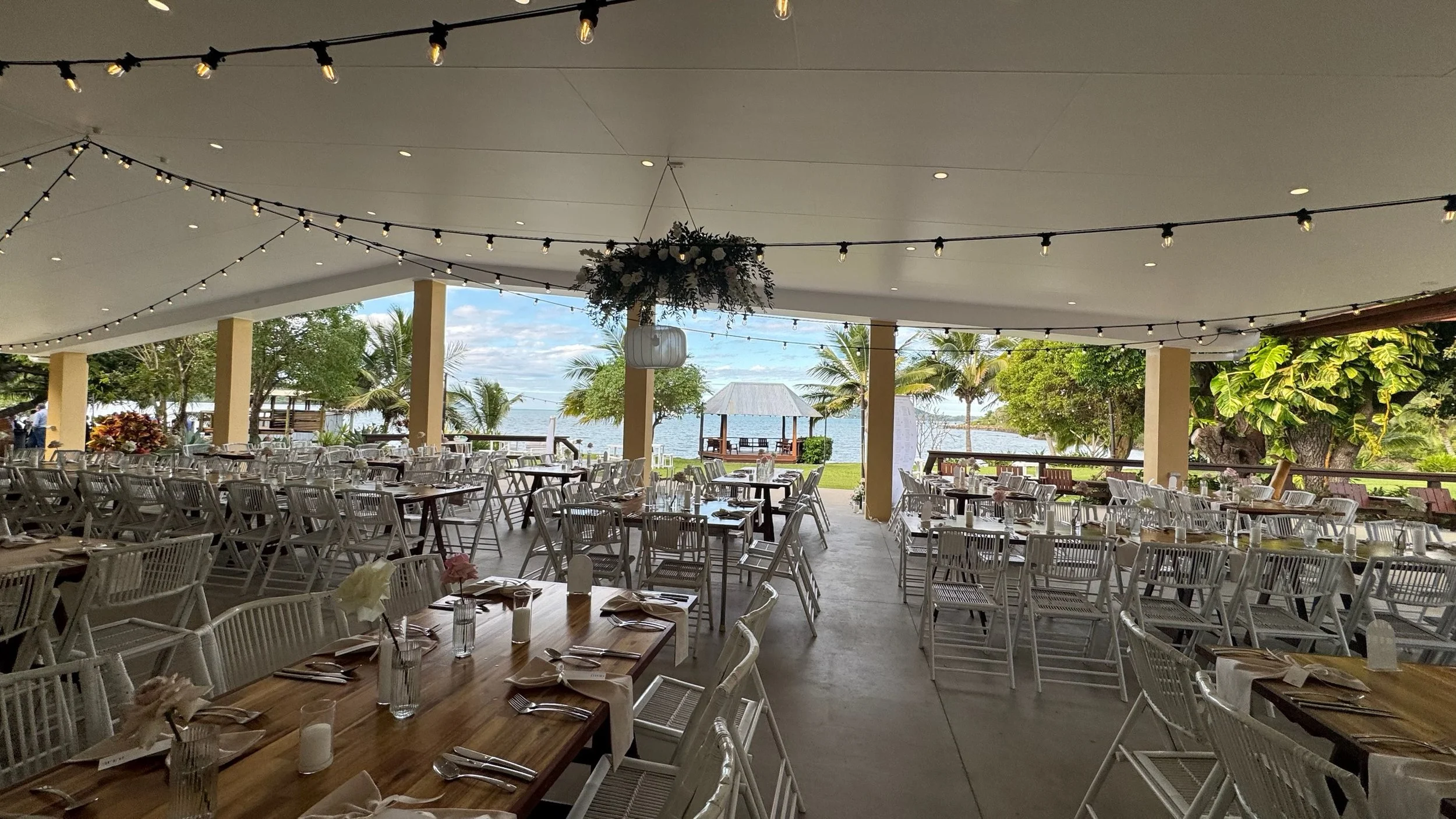 Open-air dining area with decorated tables, surrounded by tropical trees and facing a waterfront with a gazebo.