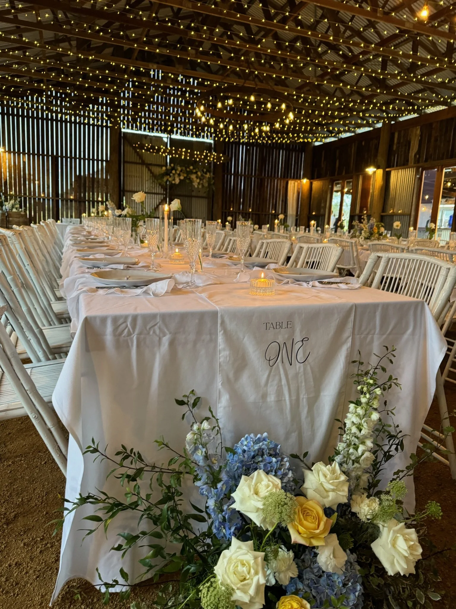A beautifully decorated wedding reception table labeled 'Table One' with white tablecloth, floral centerpiece, glassware, and candles. The venue has string lights hanging from the ceiling, wooden walls, and windows, creating a warm, festive atmospher