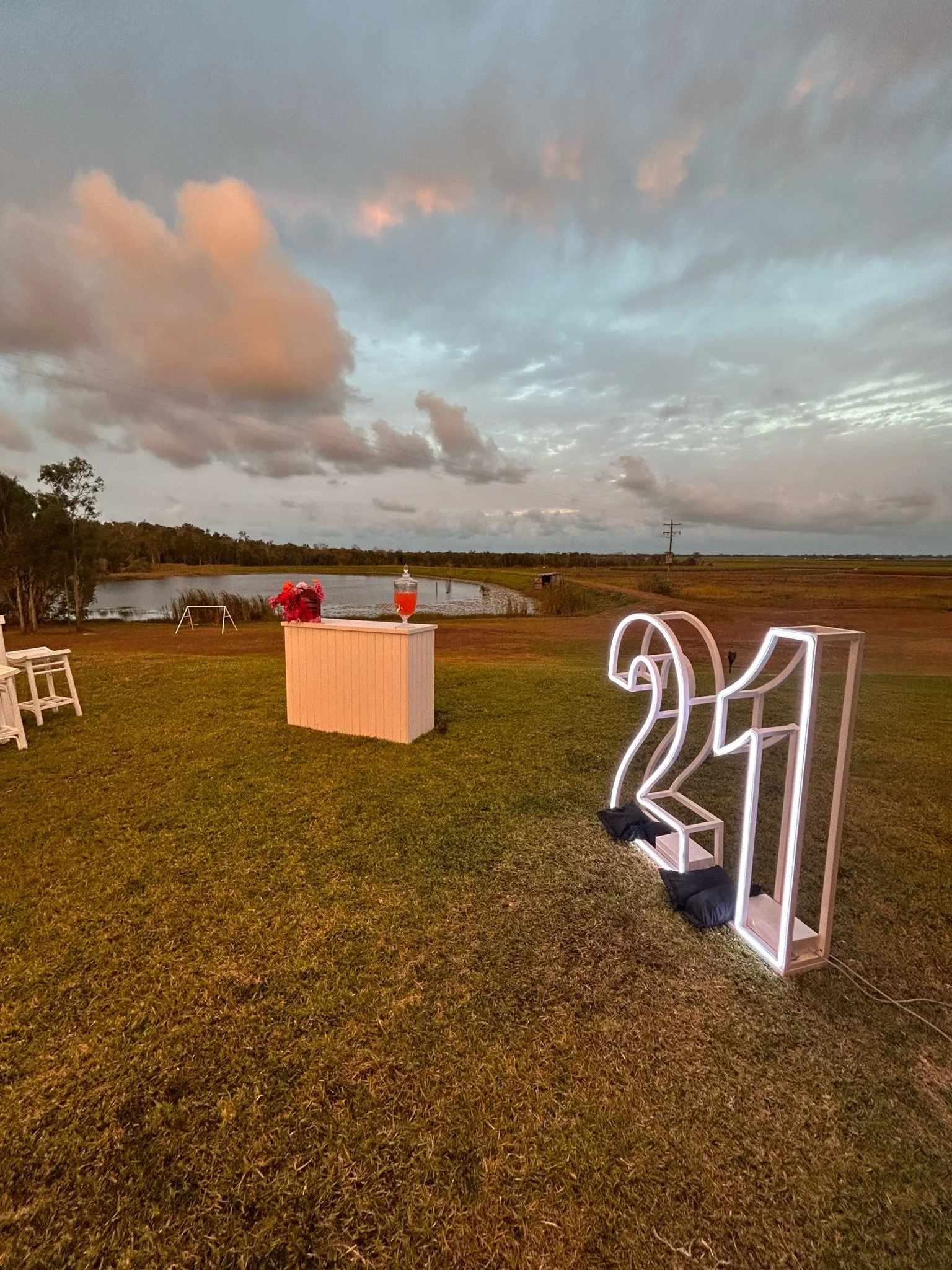 Outdoor celebration setup with a neon '21' sign on green grass, a white table with a bouquet of pink flowers and a drink, and a scenic backdrop of a river, trees, and cloudy sky during sunset.
