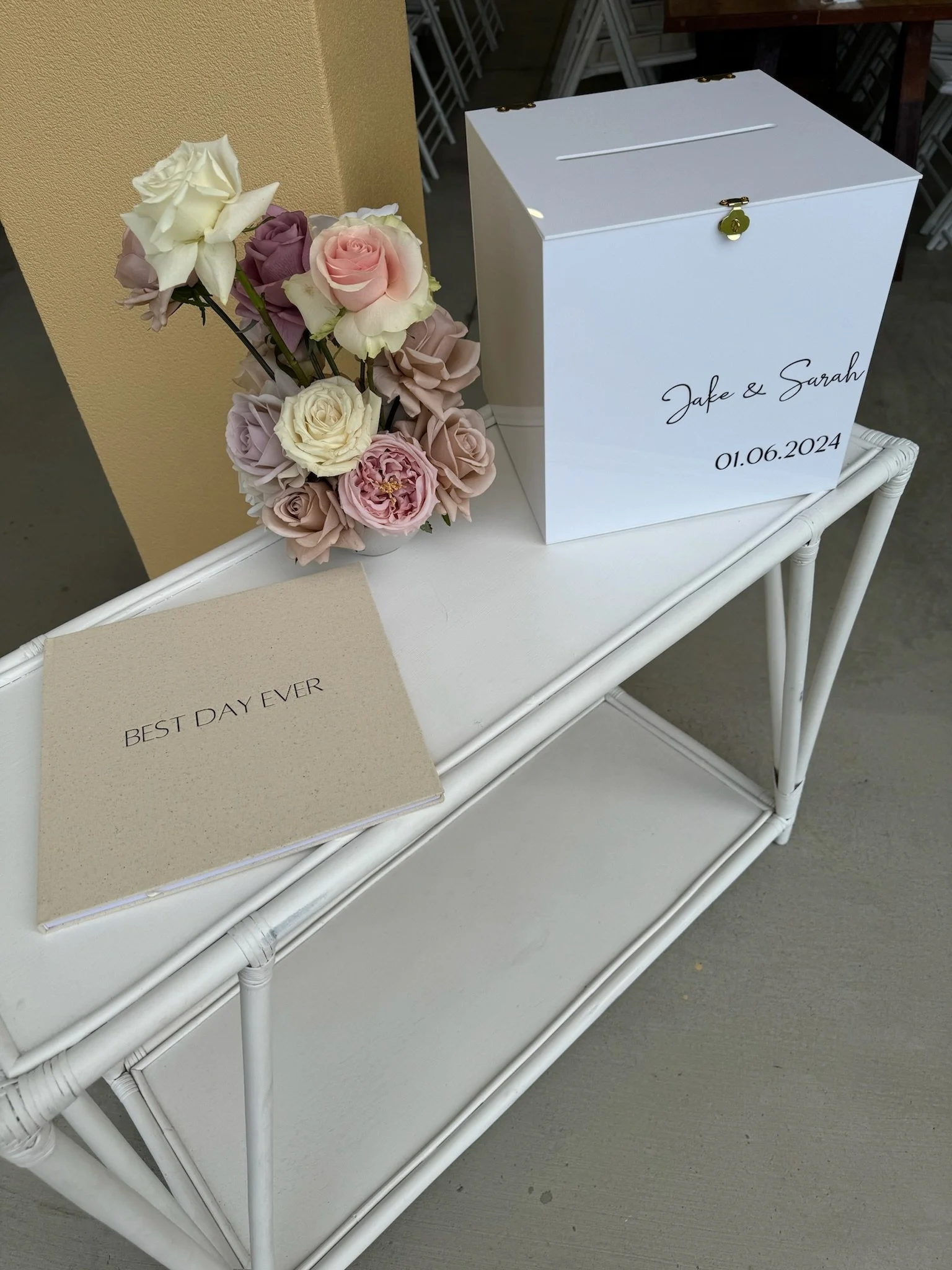 White wedding donation box with the names Jake and Sarah and the date 01.06.2024. A bouquet of pastel-colored roses and peonies and a card that reads "Best Day Ever" are placed beside it.