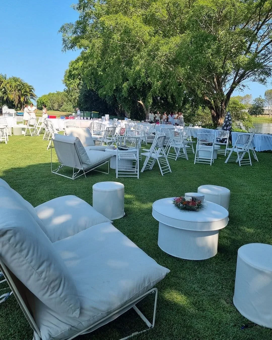White outdoor furniture including chairs, sofas, and coffee tables set up on grass for a gathering, with large trees in the background and a lake nearby.