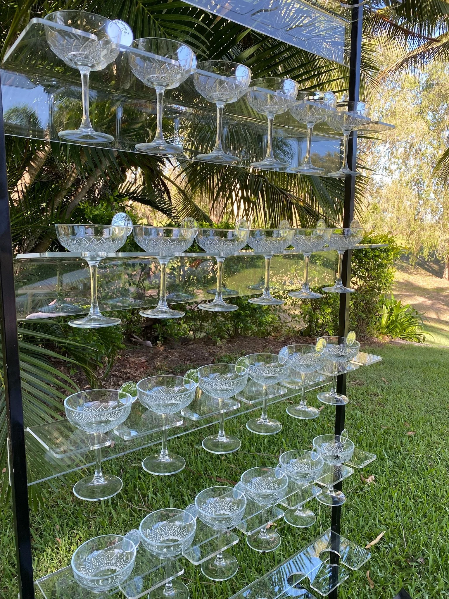 A black metal and clear acrylic shelf holds multiple empty crystal glasses and bowls with intricate patterns, set outdoors amidst green trees and grass.