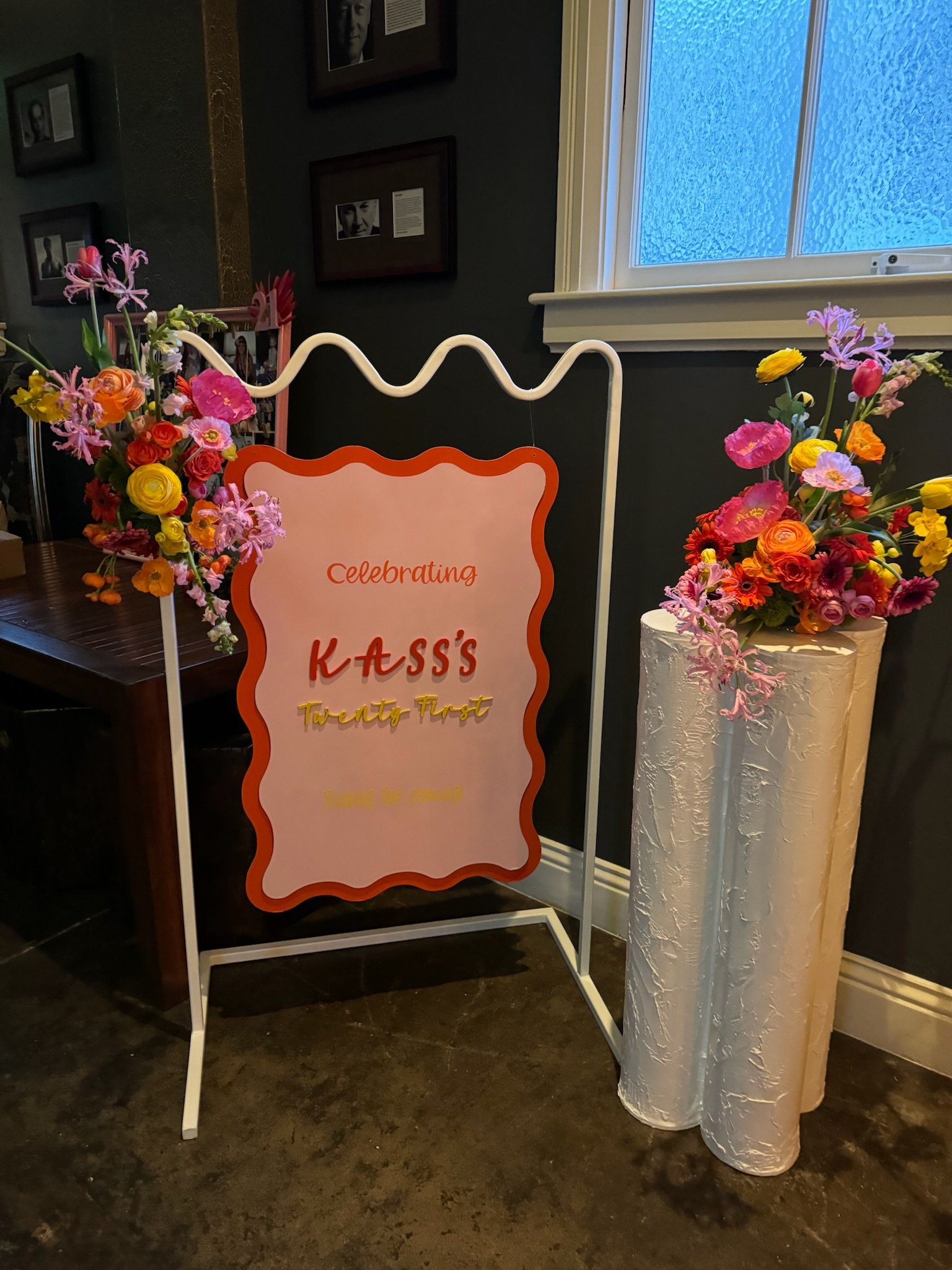 Decorative sign celebrating KASS's twenty-first birthday, surrounded by two large colorful flower arrangements in white vases, placed on a dark floor near a window and dark wall with framed pictures.