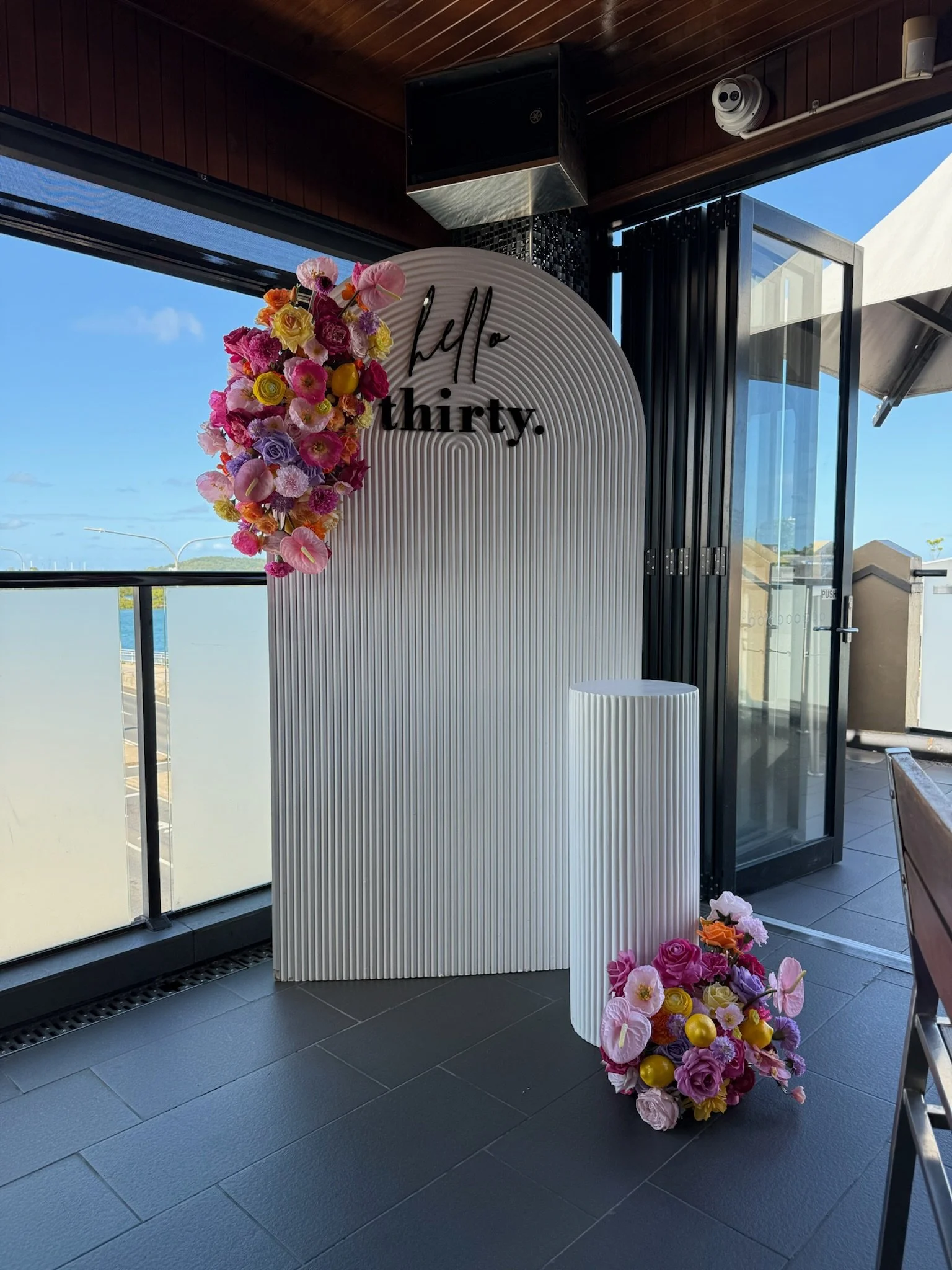 Decorative floral wedding display with a white arched backdrop and the words 'hello thirty.' written on it, featuring two arrangements of colorful flowers and gold ornaments, placed in a bright indoor space near large windows and an open glass door.