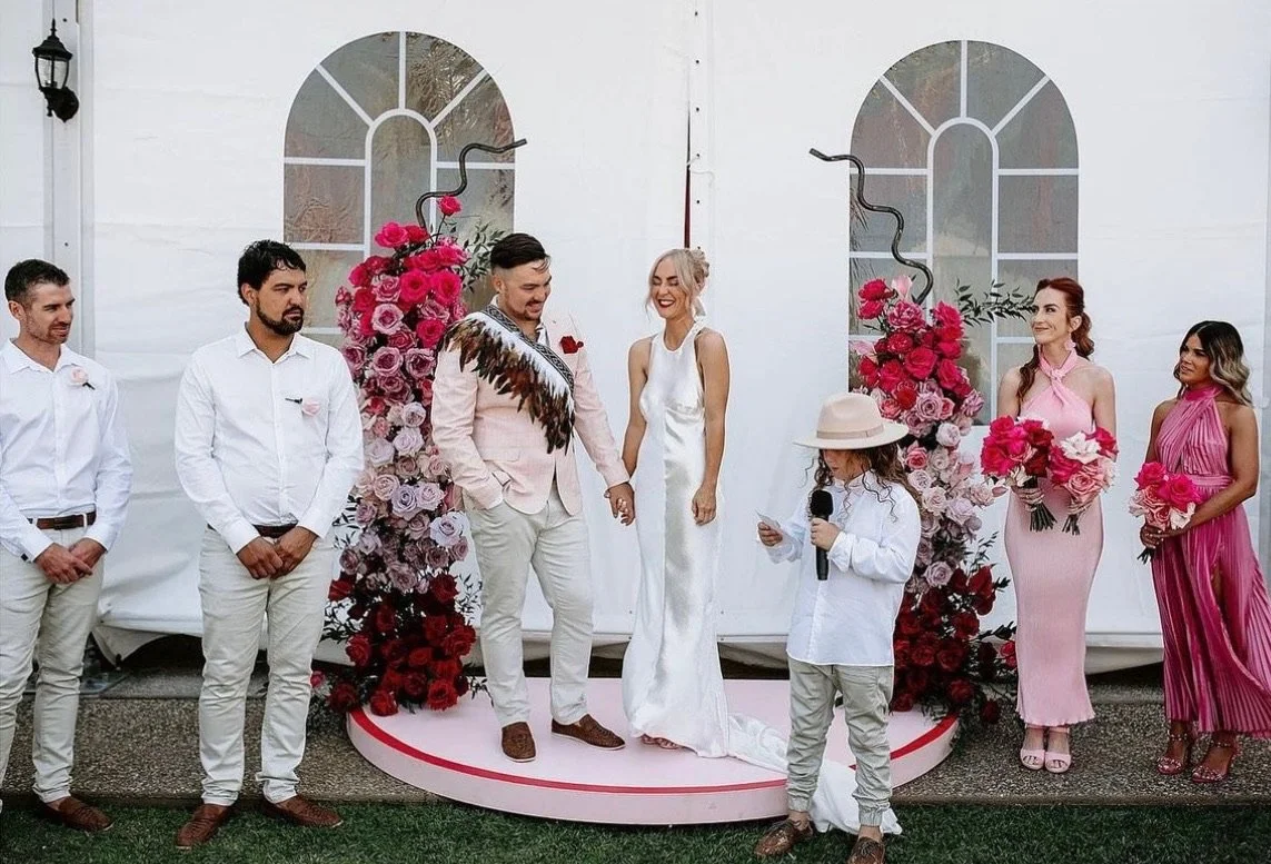 A wedding ceremony with a bride and groom holding hands, surrounded by bridesmaids and groomsmen, with a floral backdrop featuring pink and red flowers.