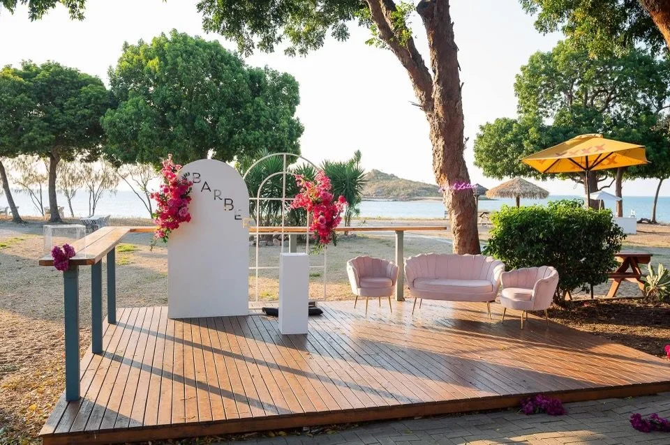 Beachside setup with a small wooden stage, pink seating, a yellow umbrella, and floral decorations, overlooking the water and surrounded by trees.