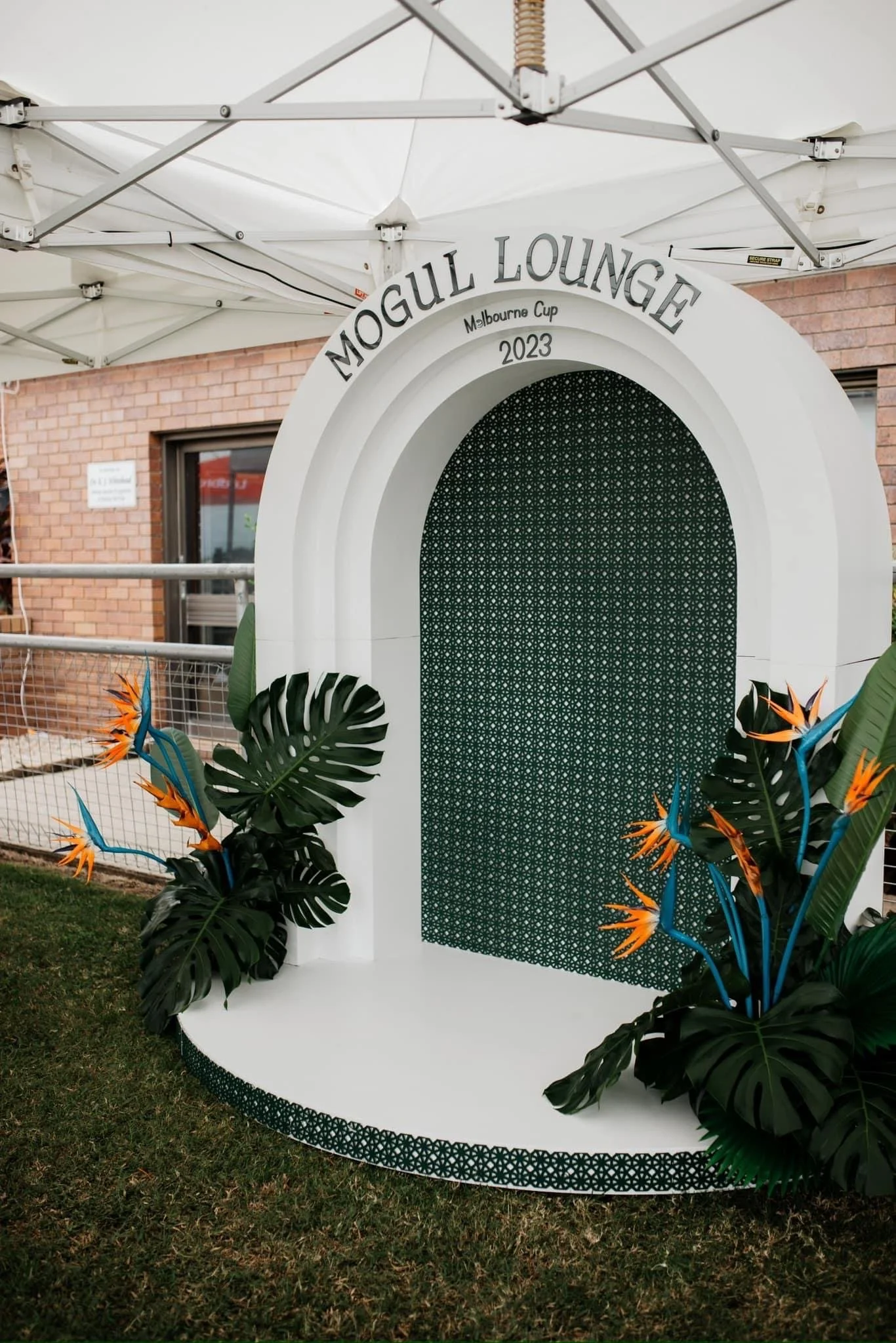 A decorative photo booth backdrop with a white arch that reads "Mogul Lounge," decorated with green patterned fabric and tropical plants with orange and blue flowers, set under a white canopy at an outdoor event.