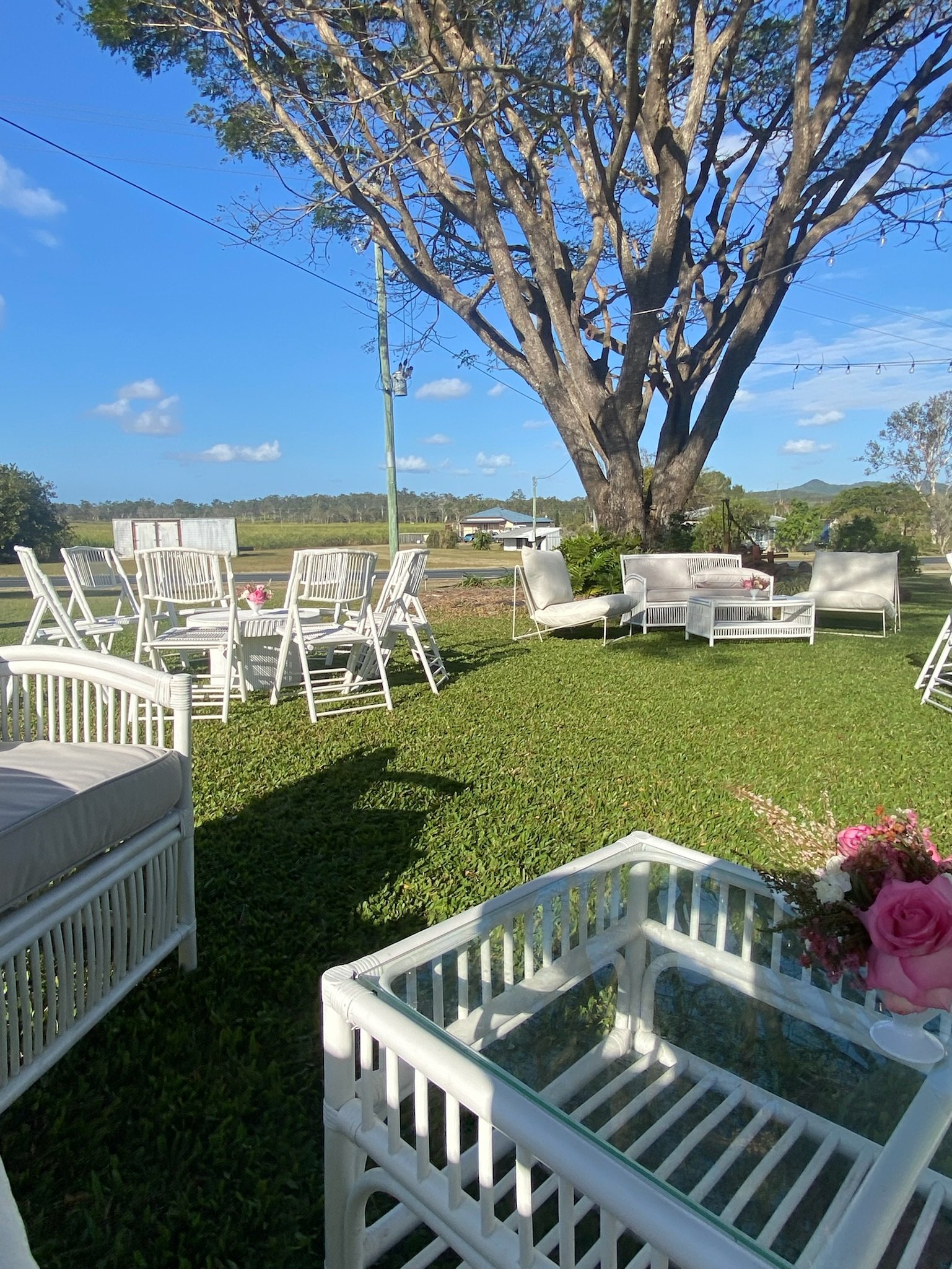 Outdoor seating area with white chairs and couches under a large tree on a grassy lawn, with blue sky and scattered clouds.
