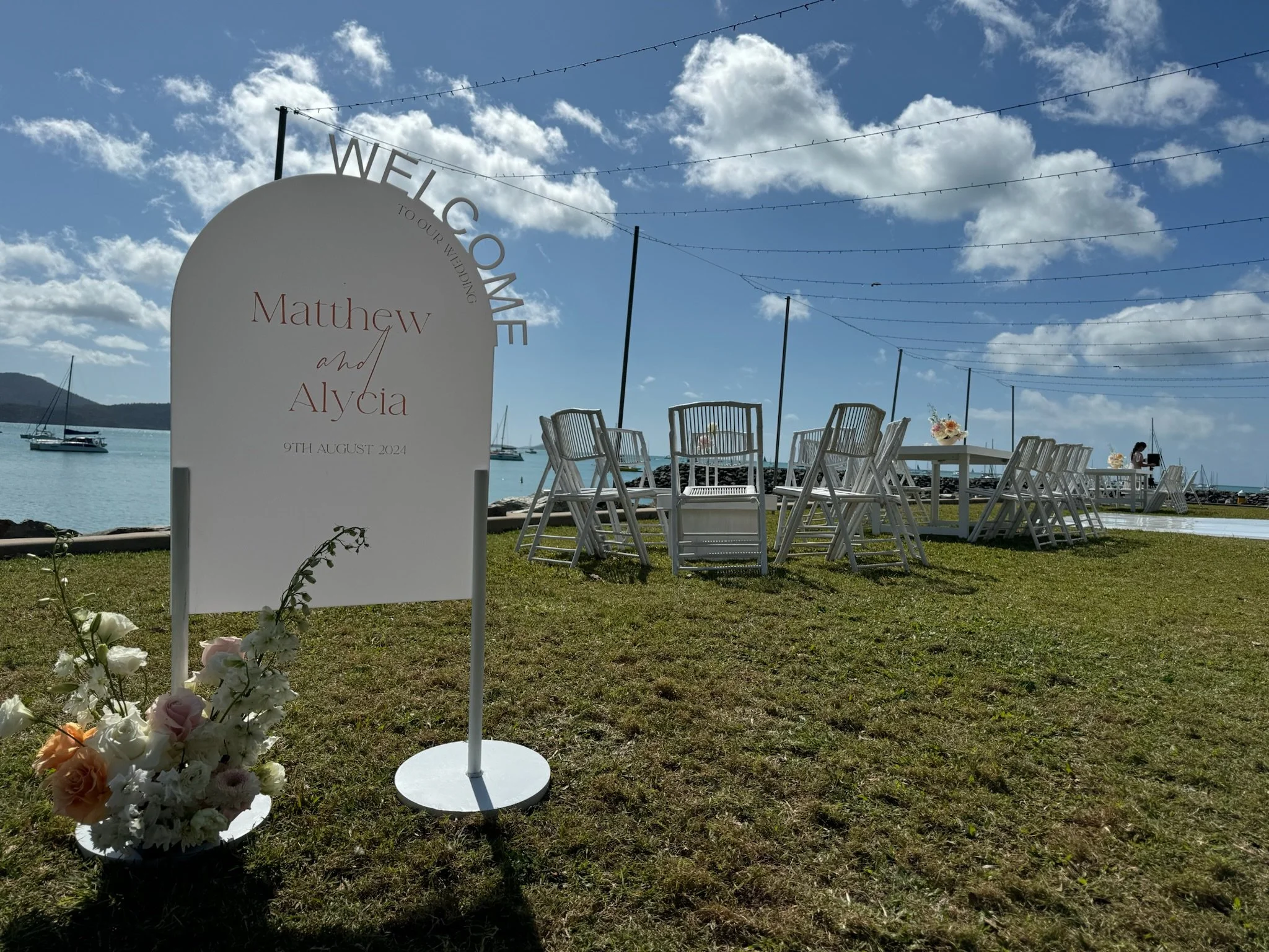 Wedding setup with outdoor tables and chairs by the water, with a wedding sign that reads 'Welcome to our wedding, Matthew and Alycia, 9th August 2024', and a floral arrangement nearby, against a background of blue sky, clouds, and sailboats.