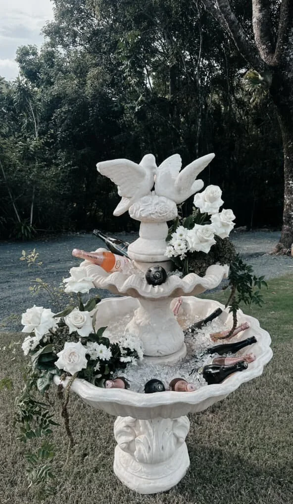 Decorative outdoor fountain with white flowers, a dove sculpture, and assorted glass bottles and candles, set in a garden with trees in the background.