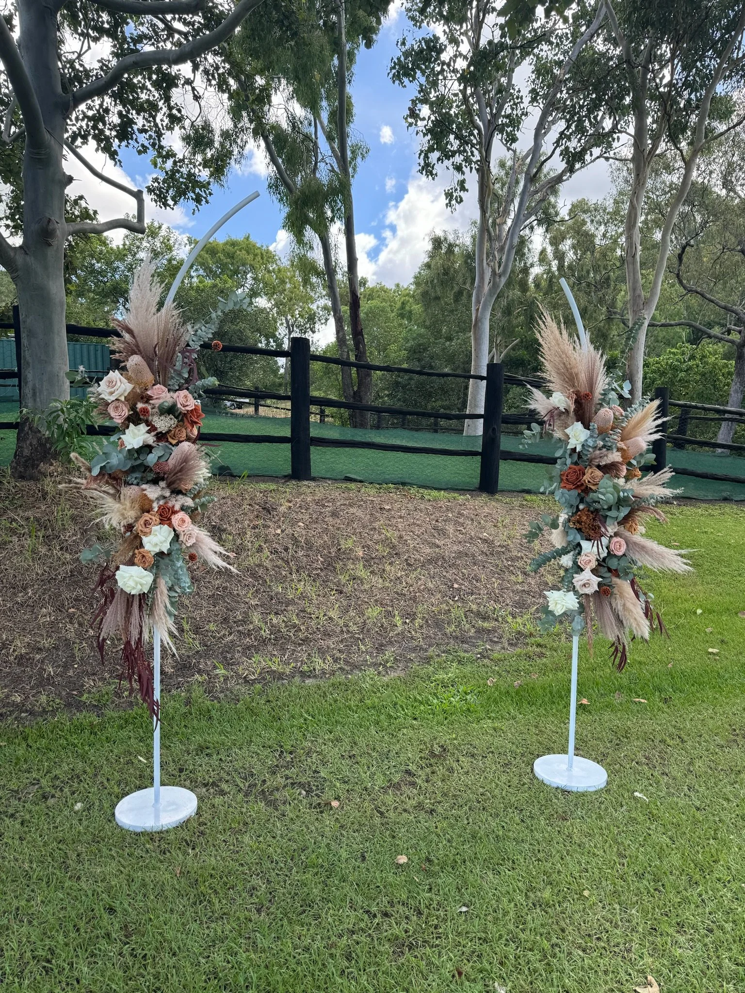 Two tall floral arrangements with roses, pampas grass, and eucalyptus on white stands outdoors in a grassy area with trees and a fence in the background.