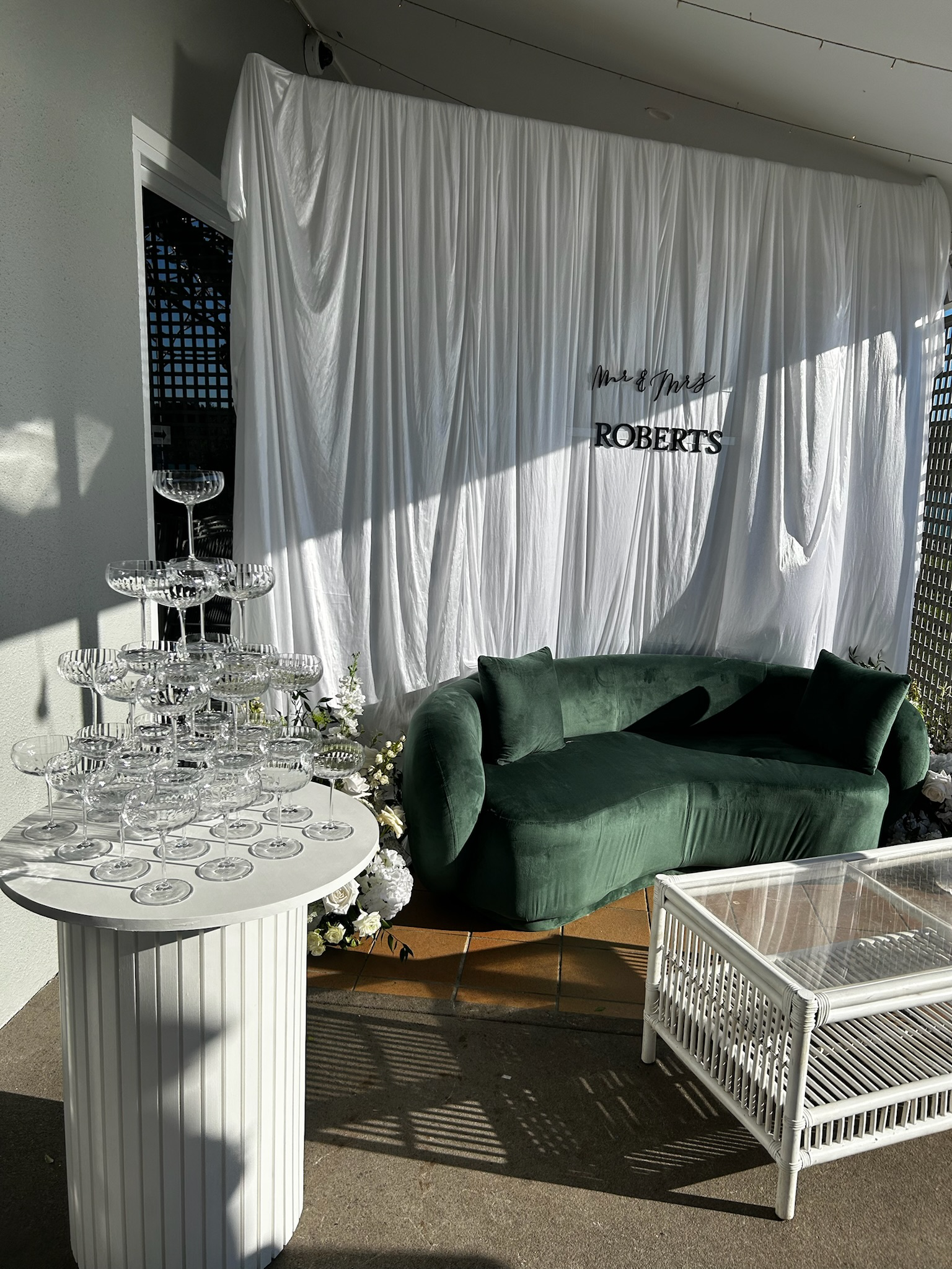 Outdoor wedding decor with a white draped backdrop that says 'Mr & Mrs Roberts,' a green velvet sofa, a white wicker table, a champagne glass tower, and floral arrangements on a patio.