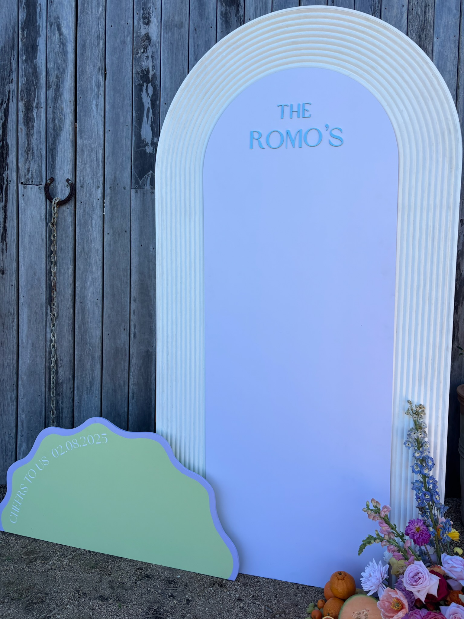 Decorative sign with white, pastel green, and purple design against a wooden fence, with flowers and small oranges at the base, and a circular sign with an arrow shape with text 'Cheers to Us 02.08.2025'.
