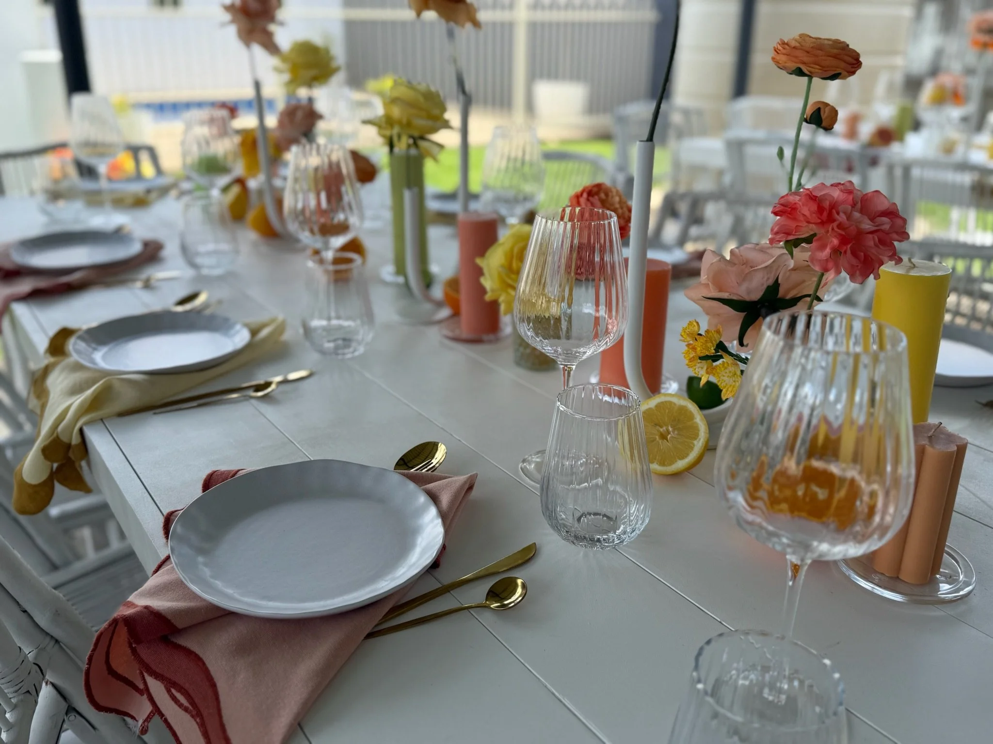 A beautifully set dining table with white tableware, gold utensils, pink and yellow cloth napkins, and glassware. The table is decorated with pink, yellow, and orange flowers in vases, and there are lemon halves and pink candles as additional decorat