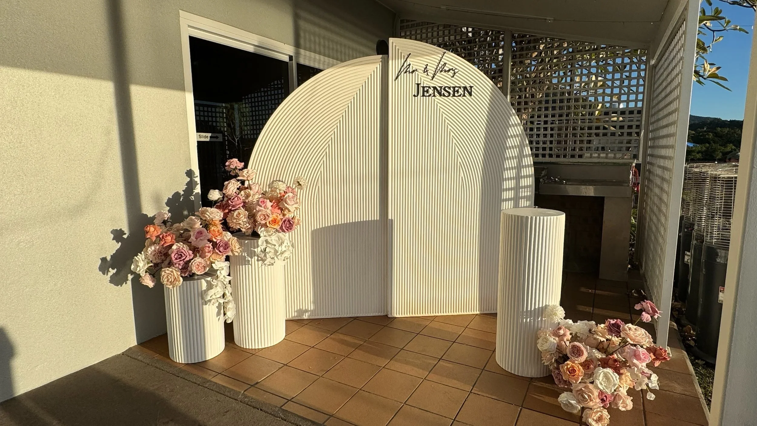 A modern outdoor celebration setup with a white folding screen, floral arrangements with pink and white roses, and a small sink on a tiled floor, with sunlight casting shadows.