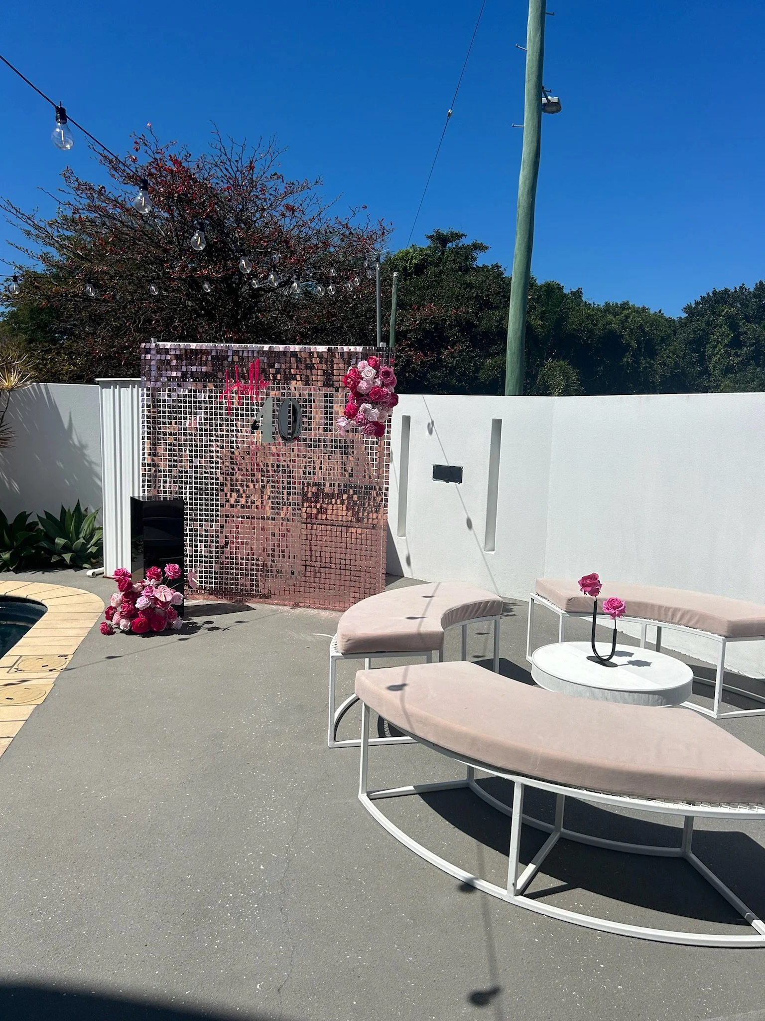 Outdoor patio decorated with pink flowers, a white wall, and a pink shimmering backdrop with the number 40 and the 'Hello' message arranged for a celebration.