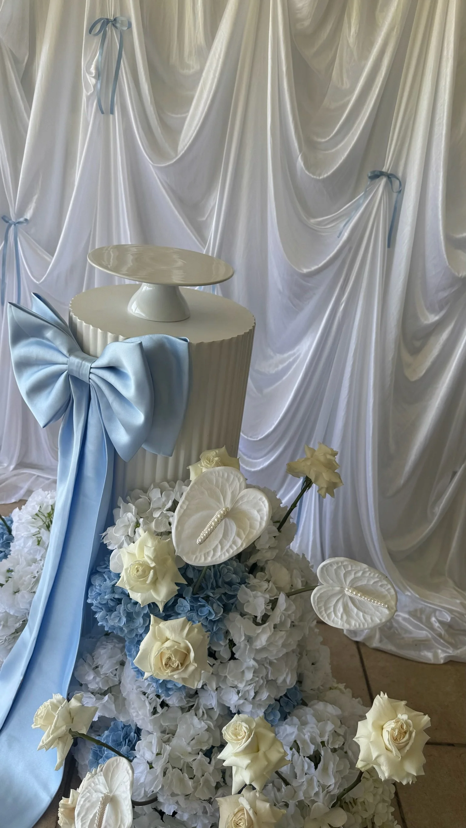 White satin backdrop with light blue ribbons, a white pedestal table with a decorative top, and a floral arrangement of white and blue flowers, including roses and anthuriums, with some small pearl accents.