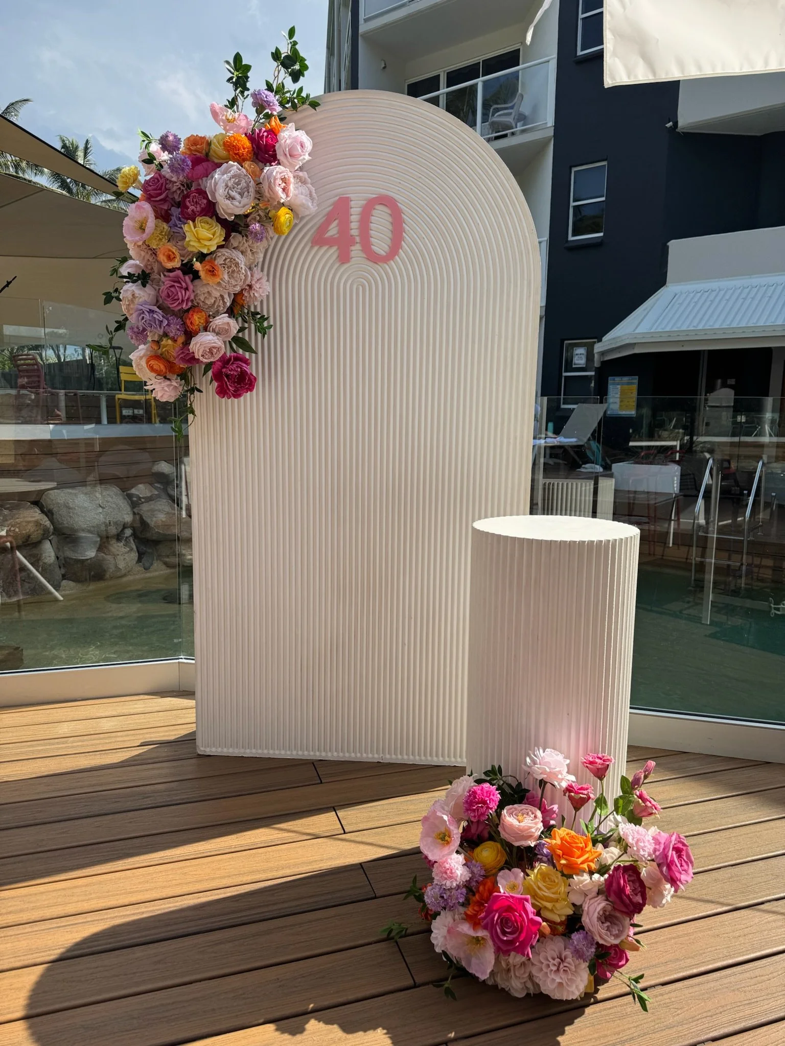 A decorative backdrop with the number 40 on it, adorned with pink, purple, yellow, orange, and white flowers, with a floral arrangement on the floor nearby, set on a wooden deck outdoor setting.