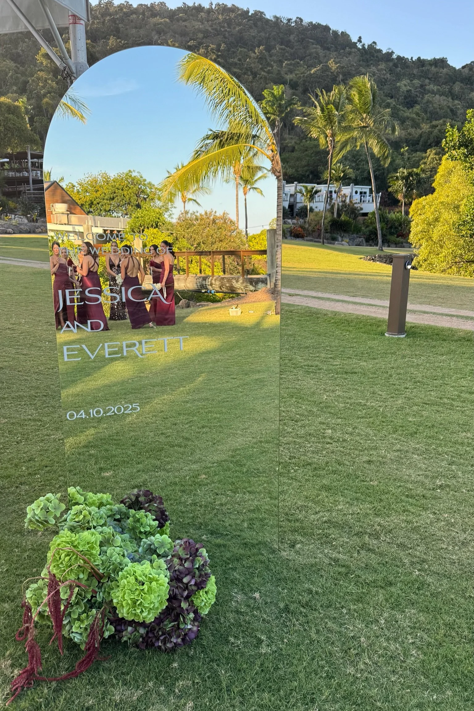 A wedding sign on a glass mirror with greenery and palm trees reflected in it, standing on a grassy area outdoors.