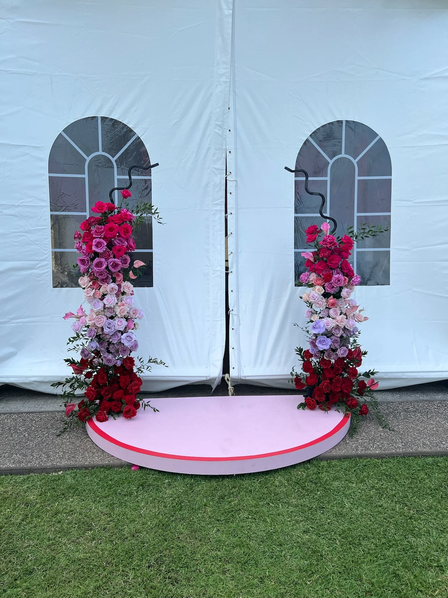 Decorative floral display with pink and red roses on black hooks in front of a white tent backdrop with window designs, and a semi-circular pink platform at the base, set on a grassy area.