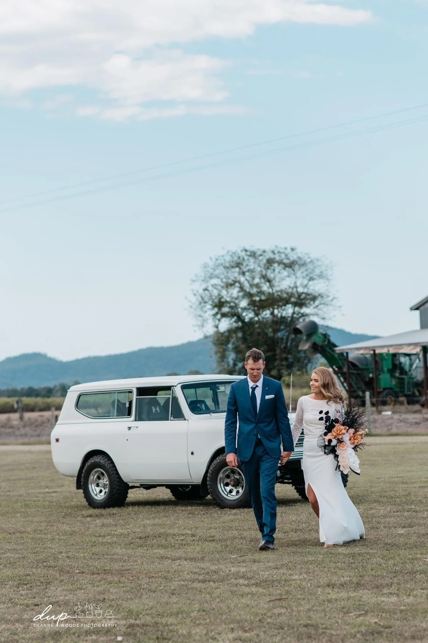 A bride and groom walking hand in hand on a grassy field, with a white vintage vehicle behind them and a rural landscape in the background.