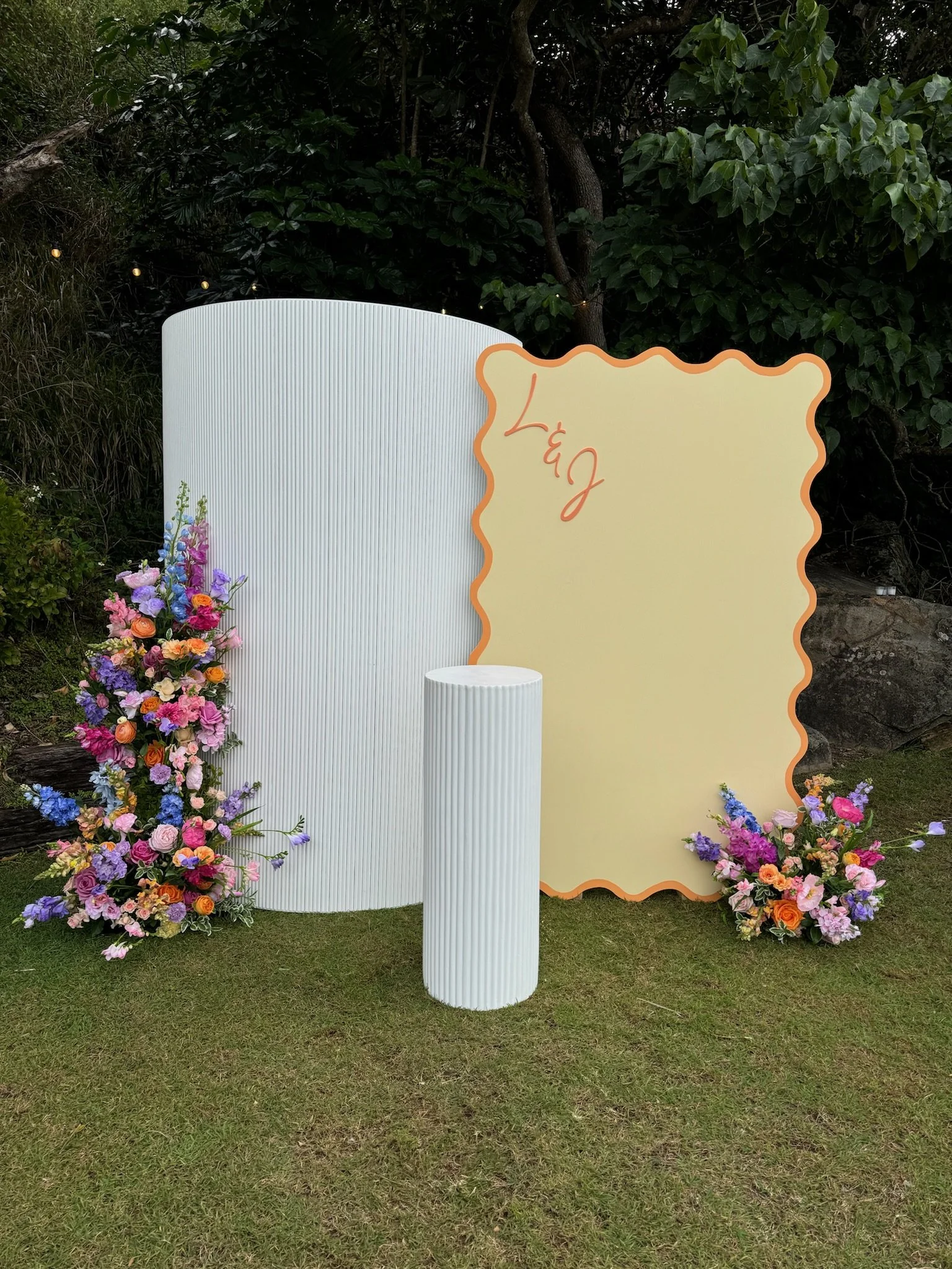A floral arrangement with pink, purple, blue, and orange flowers on the ground, a white textured backdrop, a yellow board with an orange border and the name 'Léa' written on it, and a white textured cylindrical pedestal on a grassy area with trees in