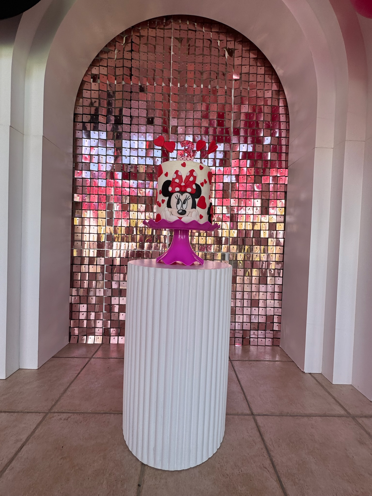 A decorative birthday cake featuring Minnie Mouse with a hat of red hearts and a pink base, placed on a white round pedestal in front of a mosaic pink and metallic backdrop.