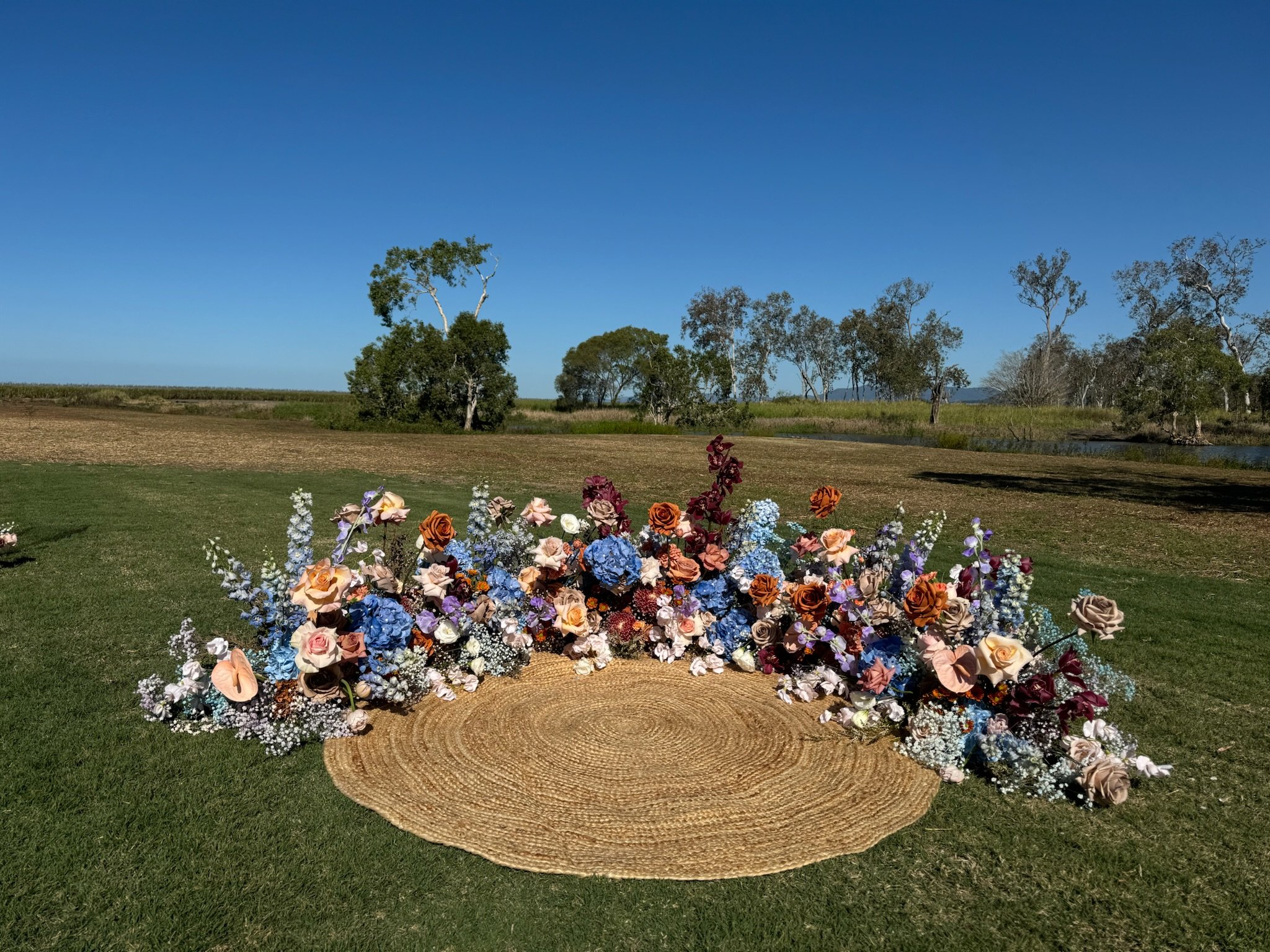 A circular woven rug on green grass, decorated with a large arrangement of colorful flowers including roses, baby's breath, and other flowers, with a rural landscape and trees in the background under a clear blue sky.