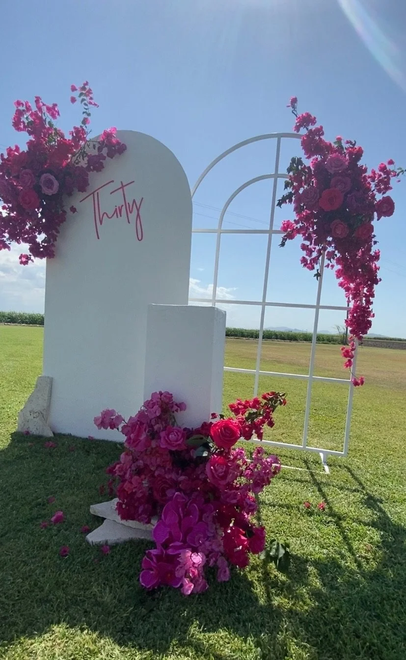 White outdoor wedding arch decorated with pink and purple flowers, with a sign that says 'Thirty' and a grassy field in the background under a blue sky.