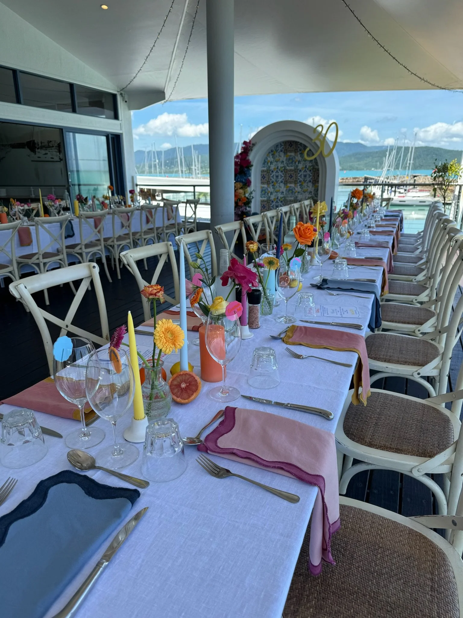 Long outdoor dining table decorated with colorful flowers, candles, and glassware, set for a celebration with a backdrop of a marina and mountains.