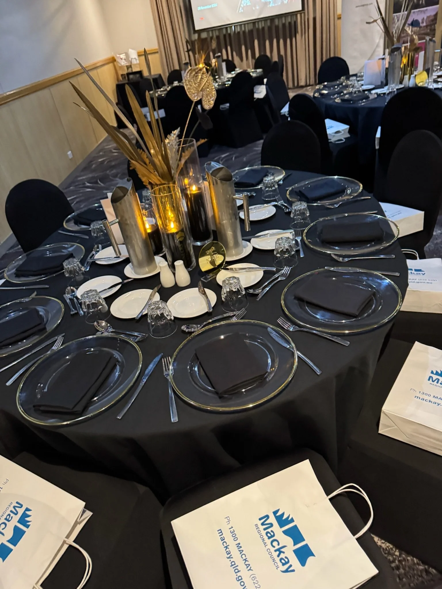 A round banquet table set for an event, with black tablecloth, black cloth napkins, wine glasses, white plates, and silverware. The centerpiece has a tall gold and black cylindrical vases with decorative gold plants and black candles. Conference fold
