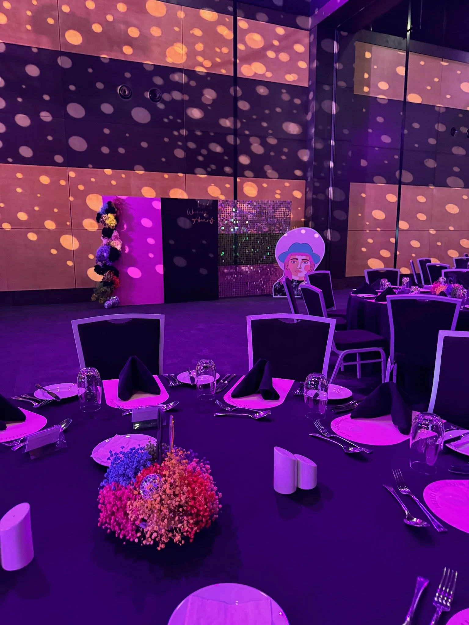 A decorated event space with round tables set for dinner, featuring black napkins, glassware, and floral centerpieces, illuminated with purple lighting and dotted light projections on the walls, with a stage area in the background.