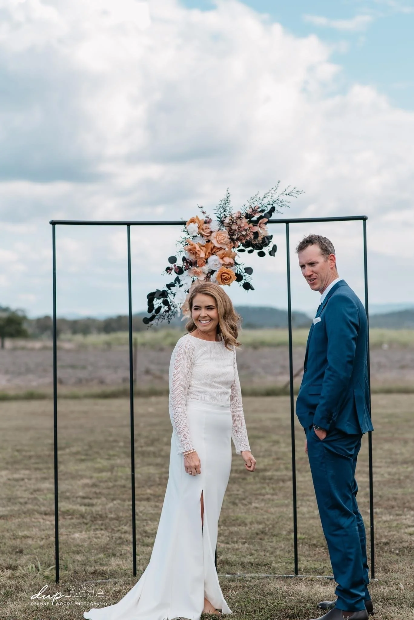 A bride and groom standing outdoors in front of a black metal arch decorated with pink, peach, and white flowers and greenery during a wedding ceremony.