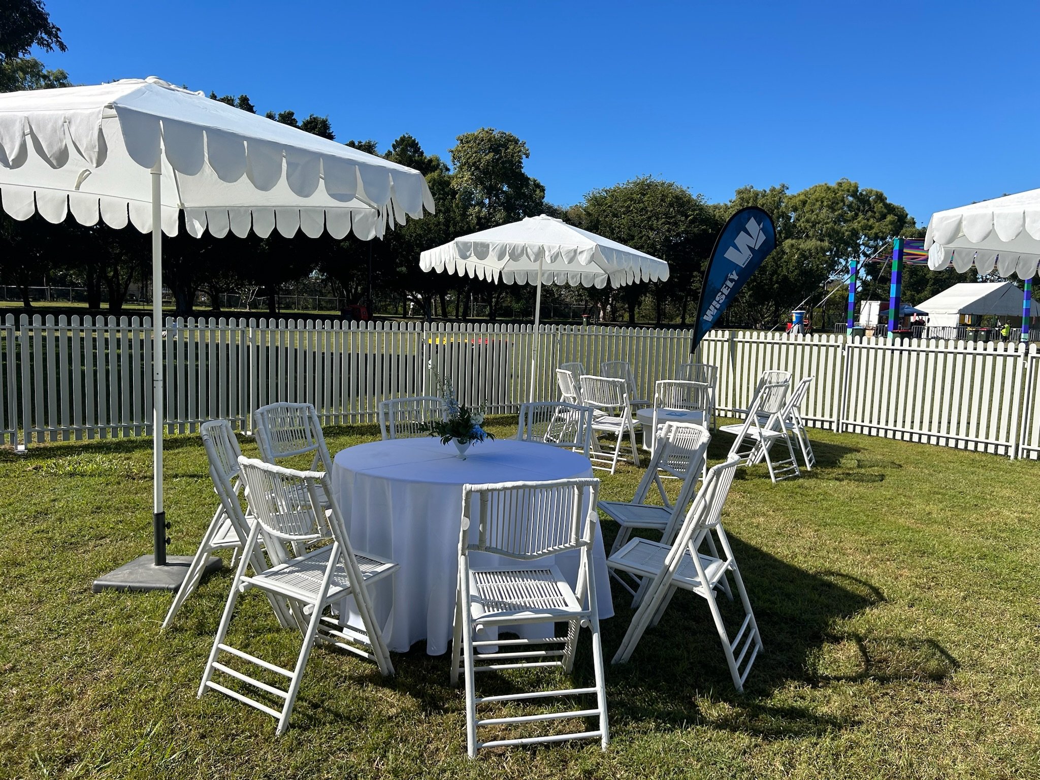 Outdoor event setup with white round table, white folding chairs, large white umbrellas, and a white picket fence on a grassy area under a clear blue sky.