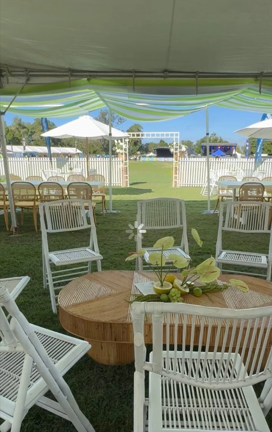 Outdoor event setup with white chairs, wooden tables, umbrellas, and a grassy area, possibly for a gathering or celebration.