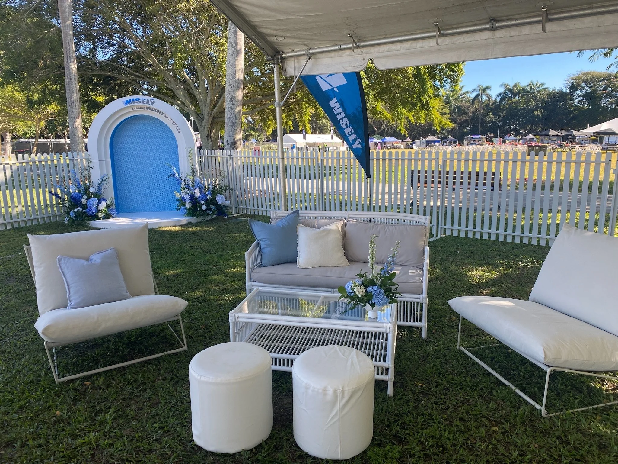 Outdoor event setup with white patio furniture, floral arrangements, a small glass table, and a decorative backdrop with blue flowers and greenery, enclosed by a white picket fence, under a large canopy with a grassy area and tents in the background.