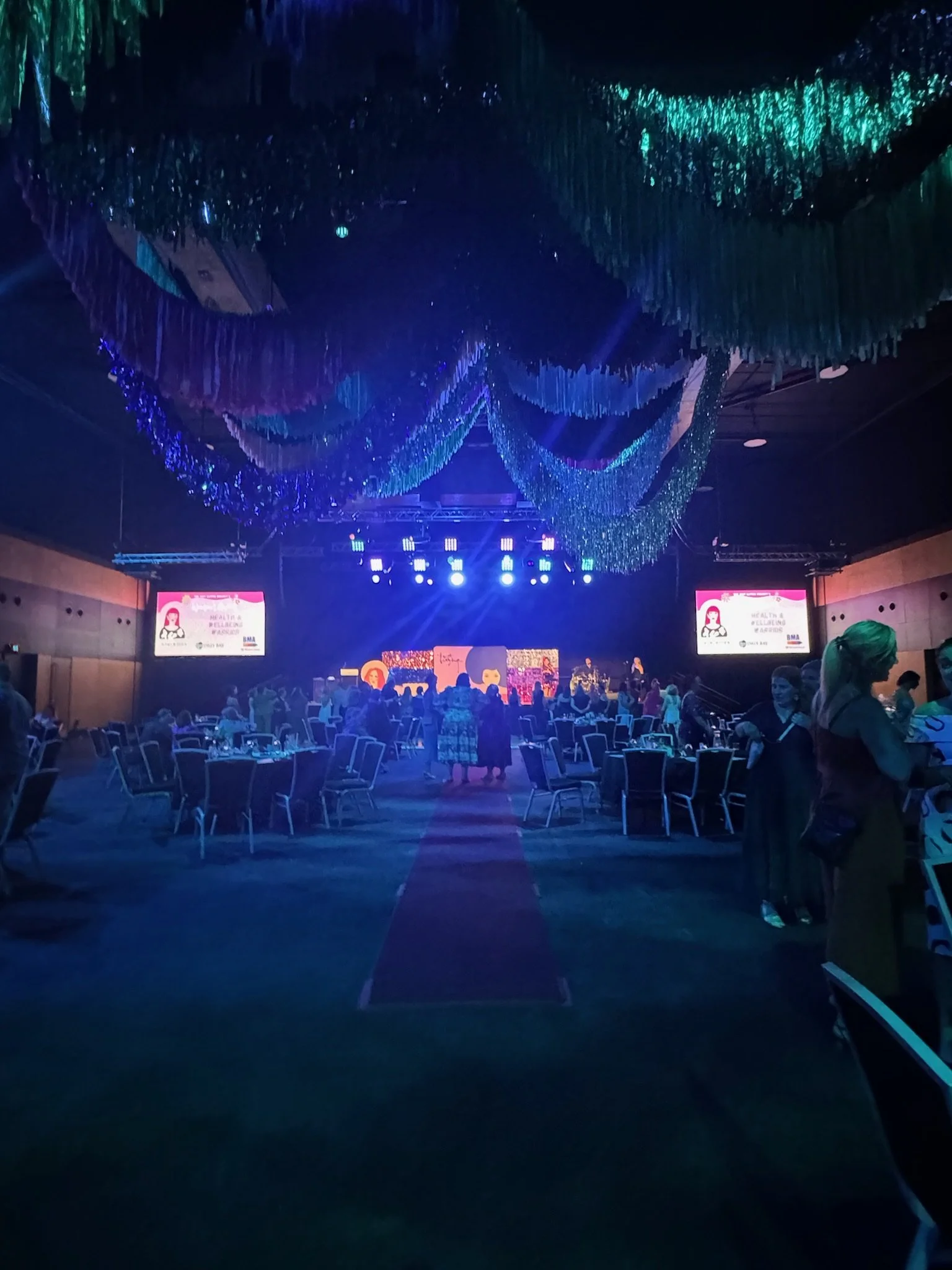 Event hall decorated with colorful streamers and lights, with a stage at the front, and round tables and chairs set up for guests.