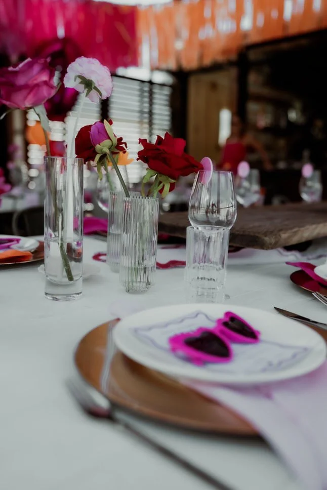 A table set for a celebration with colorful flowers in vases, place settings with napkins and plates, and pink sunglasses on a plate. The background shows a rustic setting with wooden walls and string lights.